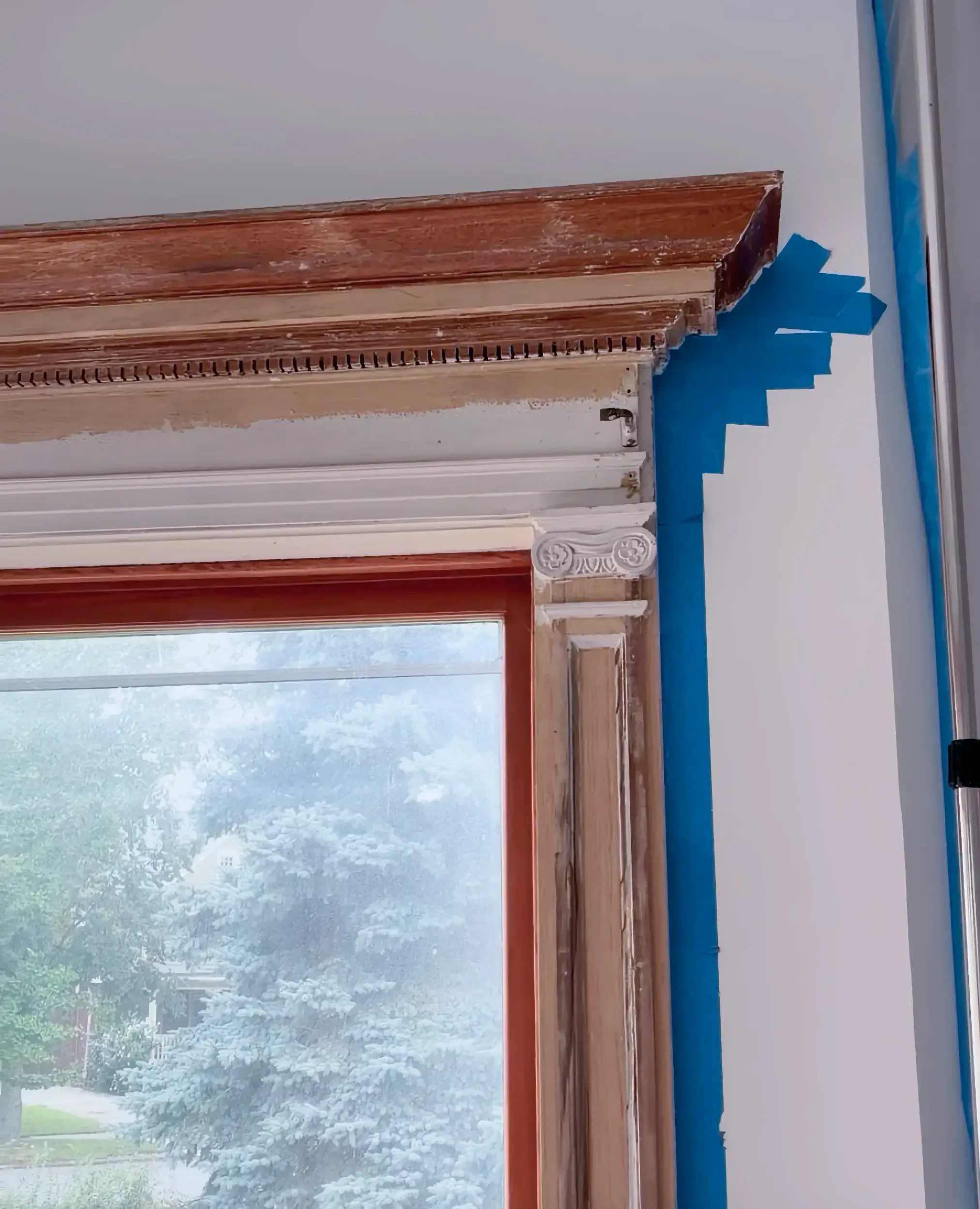 Interior of a room under renovation with a partially removed wooden door frame, blue painter's tape on the adjacent wall, and a window showing outside greenery.