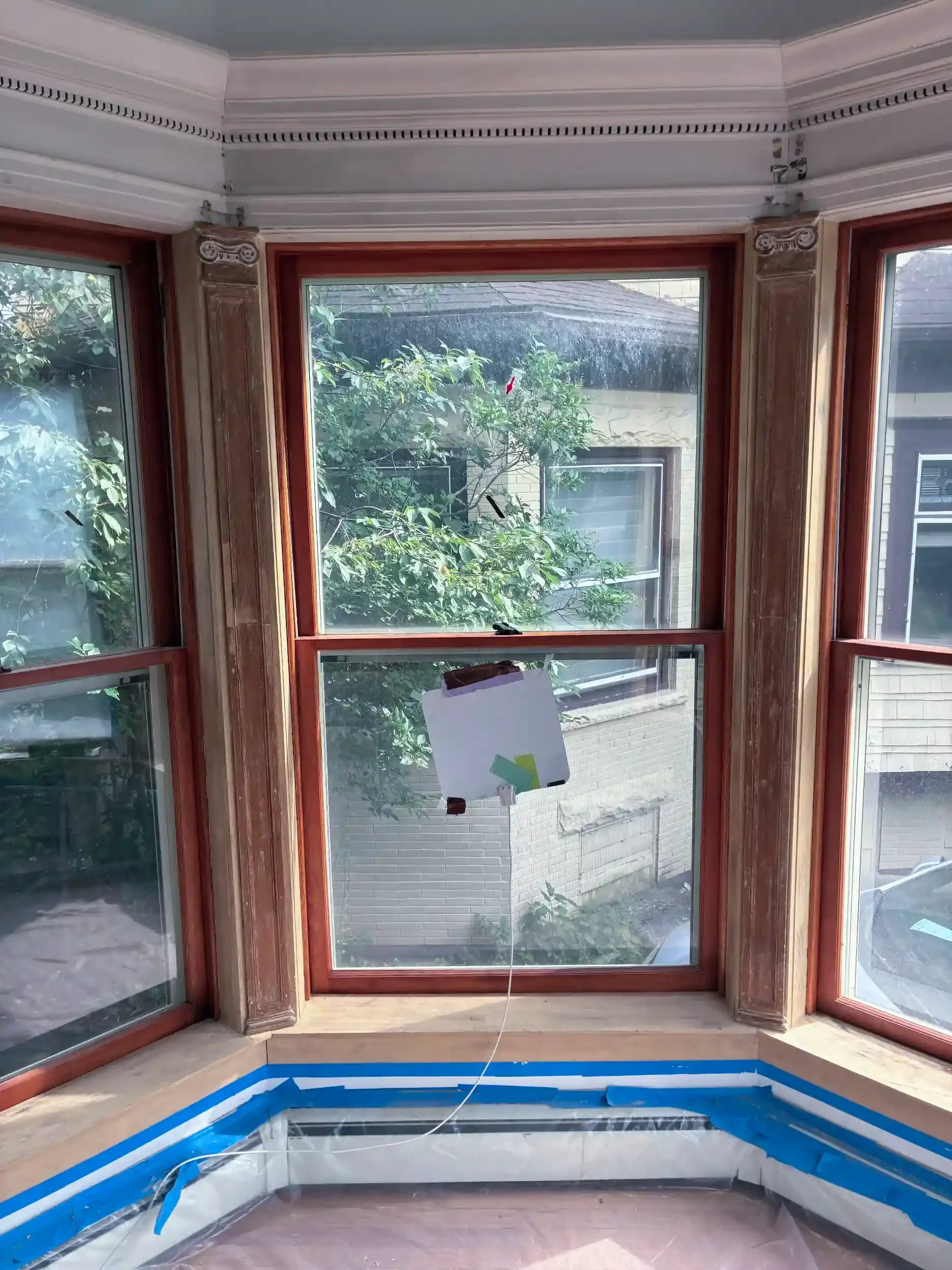 The image shows a room with large windows and a view of trees outside. The windows have wooden frames, and the interior has crown molding on the ceiling. There is a piece of paper attached to a string hanging down, and a blue painter's tape is applied along the bottom edge of the window frame.
