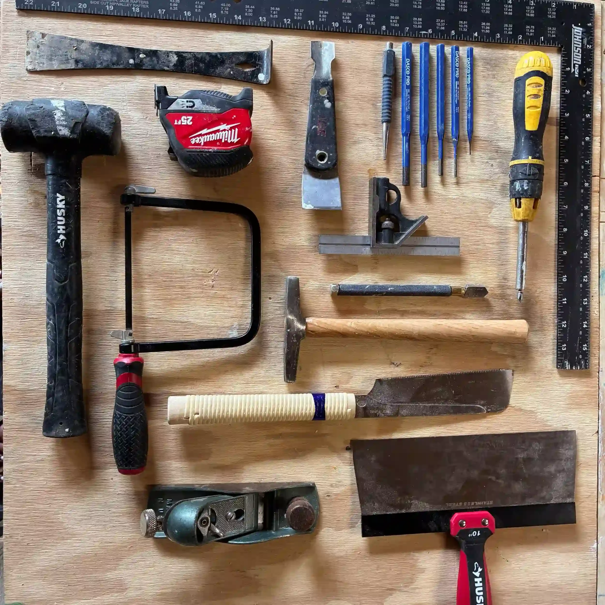 Collection of various hand tools including a hammer, tape measure, screwdrivers, chisels, a hacksaw, a spirit level, and other tools on a wooden surface, with a measuring ruler along the top and right edges of the image.
