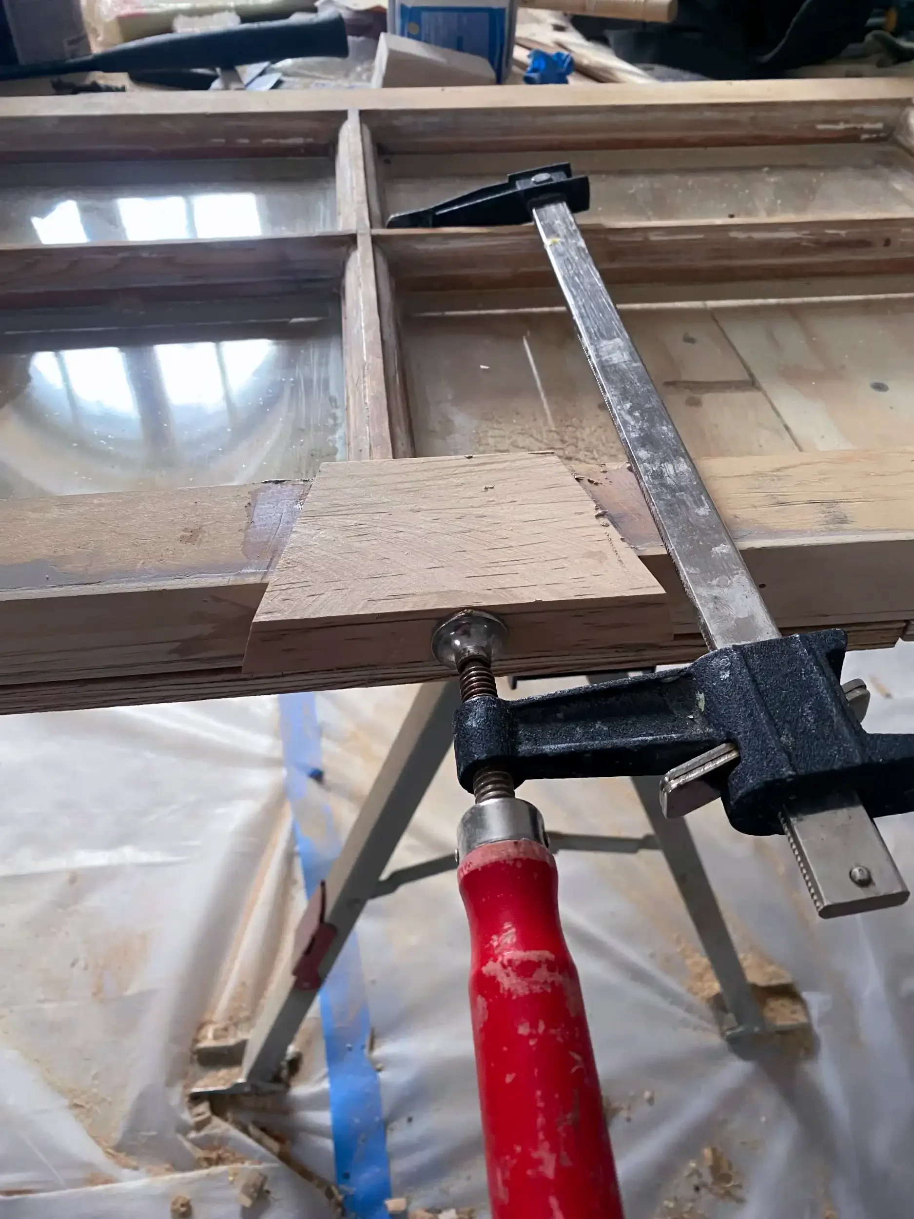 A workshop or woodworking space with a partially assembled wooden frame, clamped to a workbench, with a red clamp visible at the bottom. Tools and materials are scattered in the background.