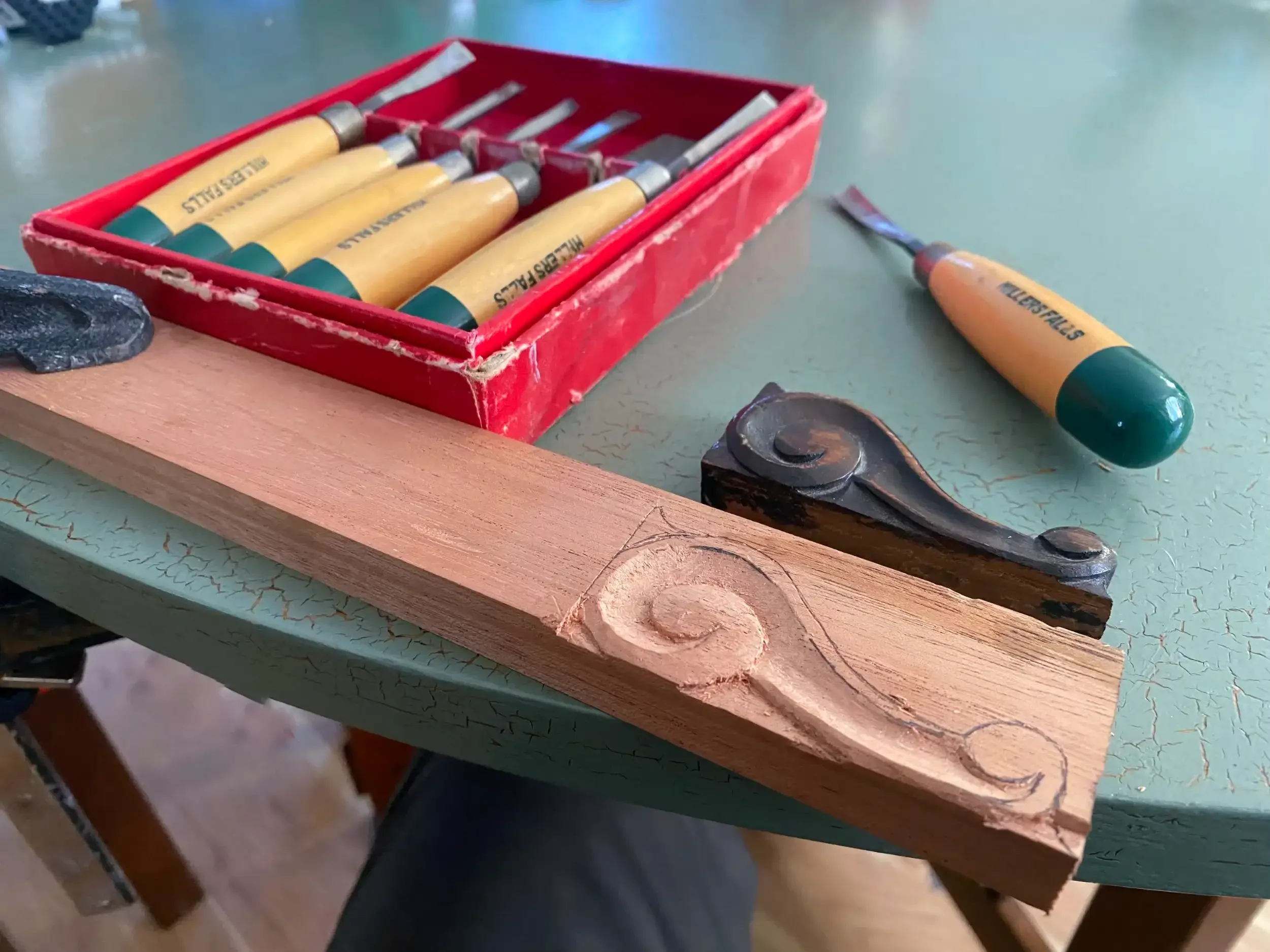 A red plastic box containing small chisels with wooden handles and metal blades, and a few small woodworking tools, set on a green surface with a textured pattern.