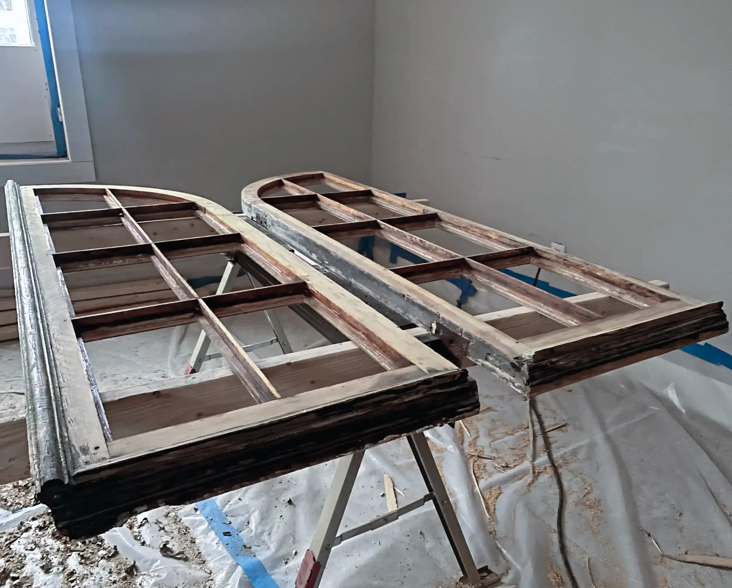 Construction site with wood frame structures on tables, walls painted gray, plastic sheets on the floor, and a window in the background.
