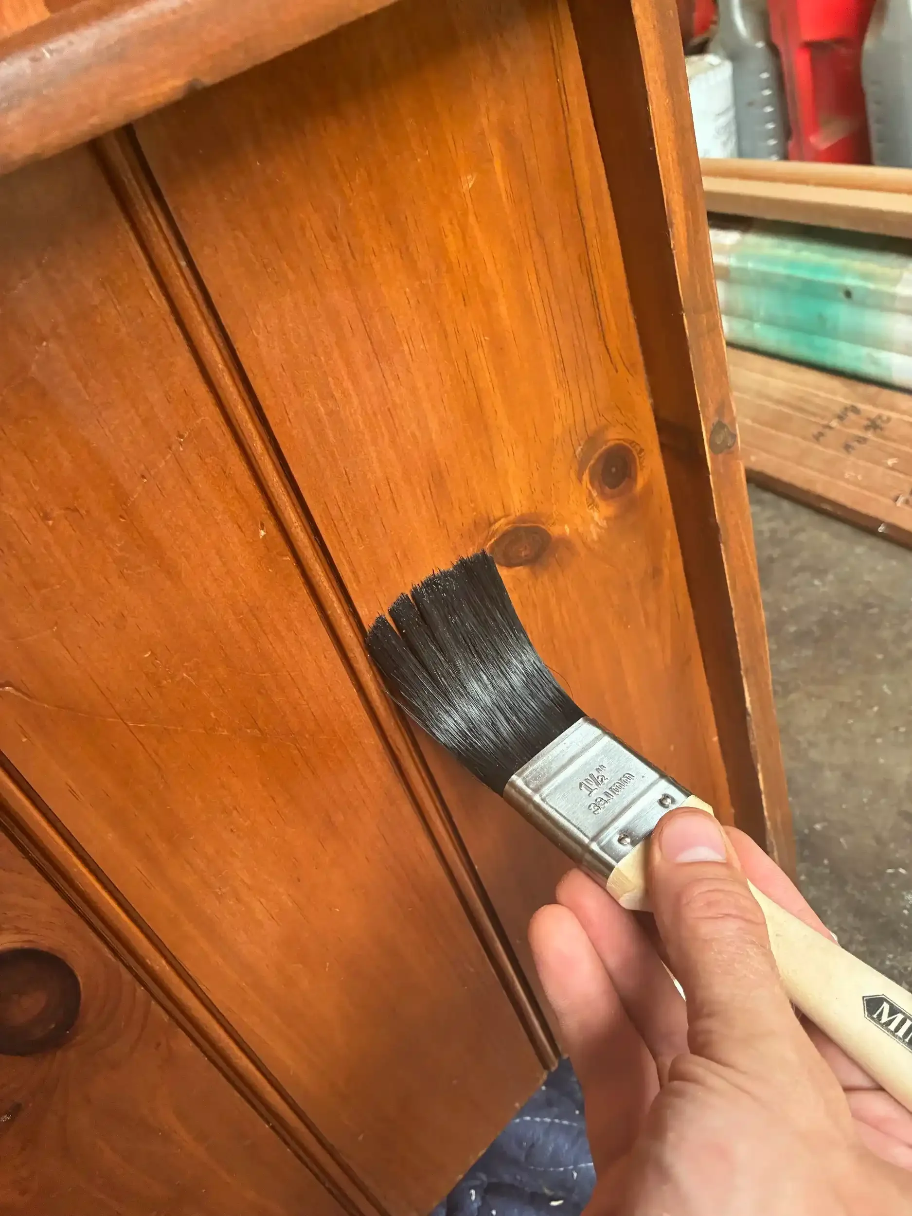 A person is painting a wooden piece of furniture with a black brush. The furniture is a warm brown color and appears to be a dresser or cabinet.