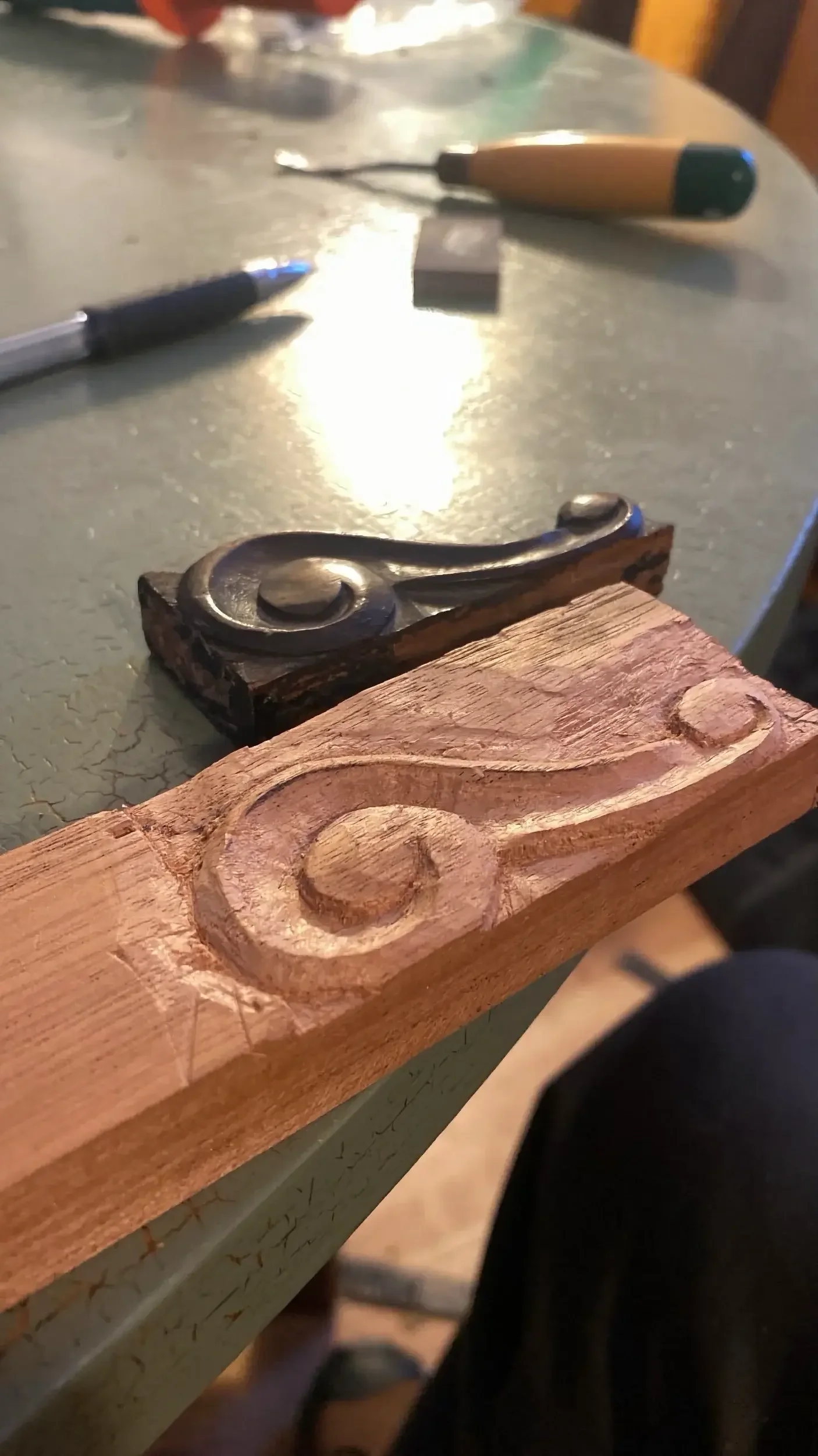 Close-up of a woodworking project in progress, showing a wooden board with carved circular indentations and a black metal fixture. Various tools are visible on a worktable in the background.
