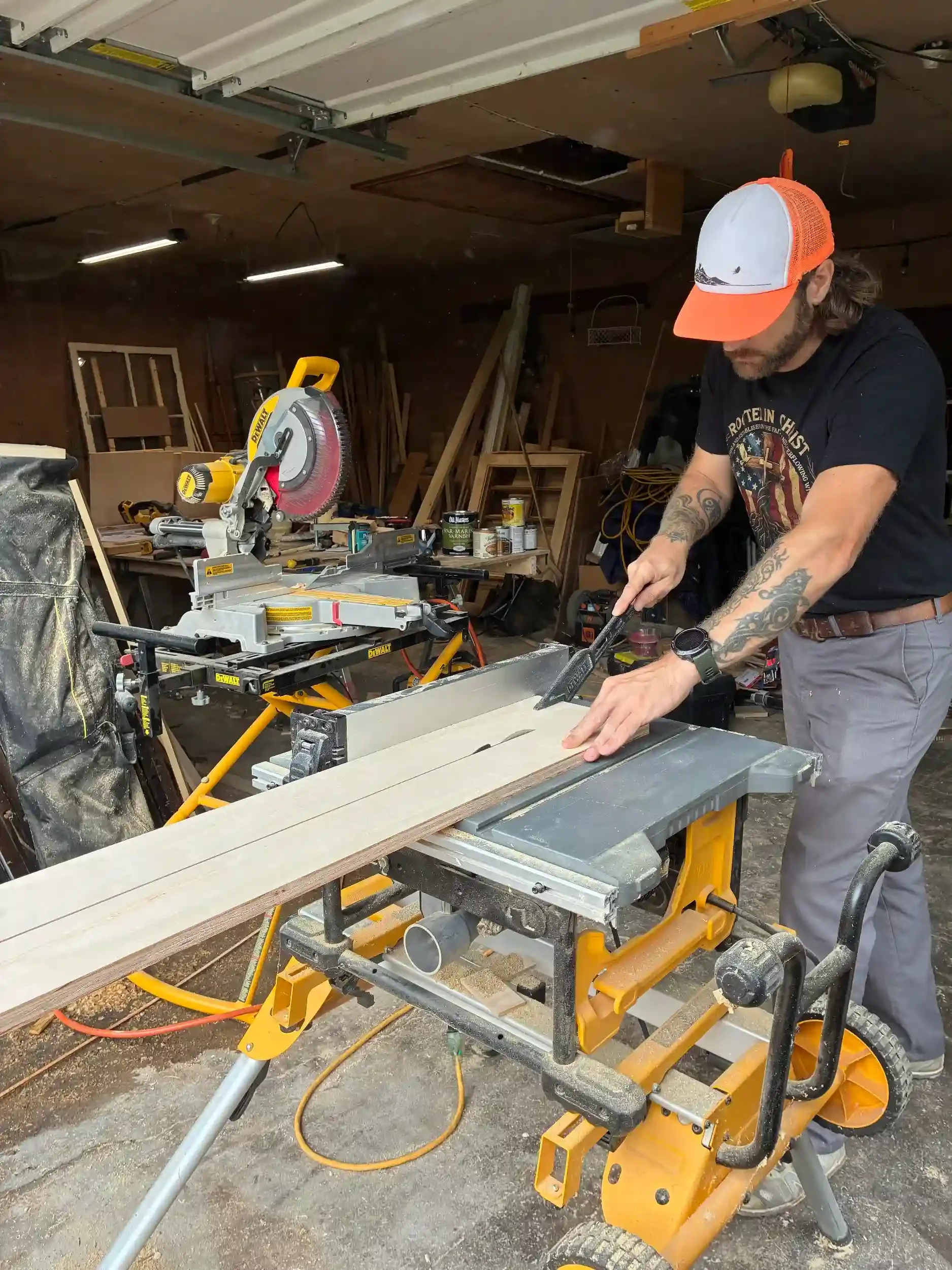 A man working with a miter saw on a wooden workbench in a woodworking workshop. The workshop has various tools, wood pieces, and supplies around, with a garage door overhead.