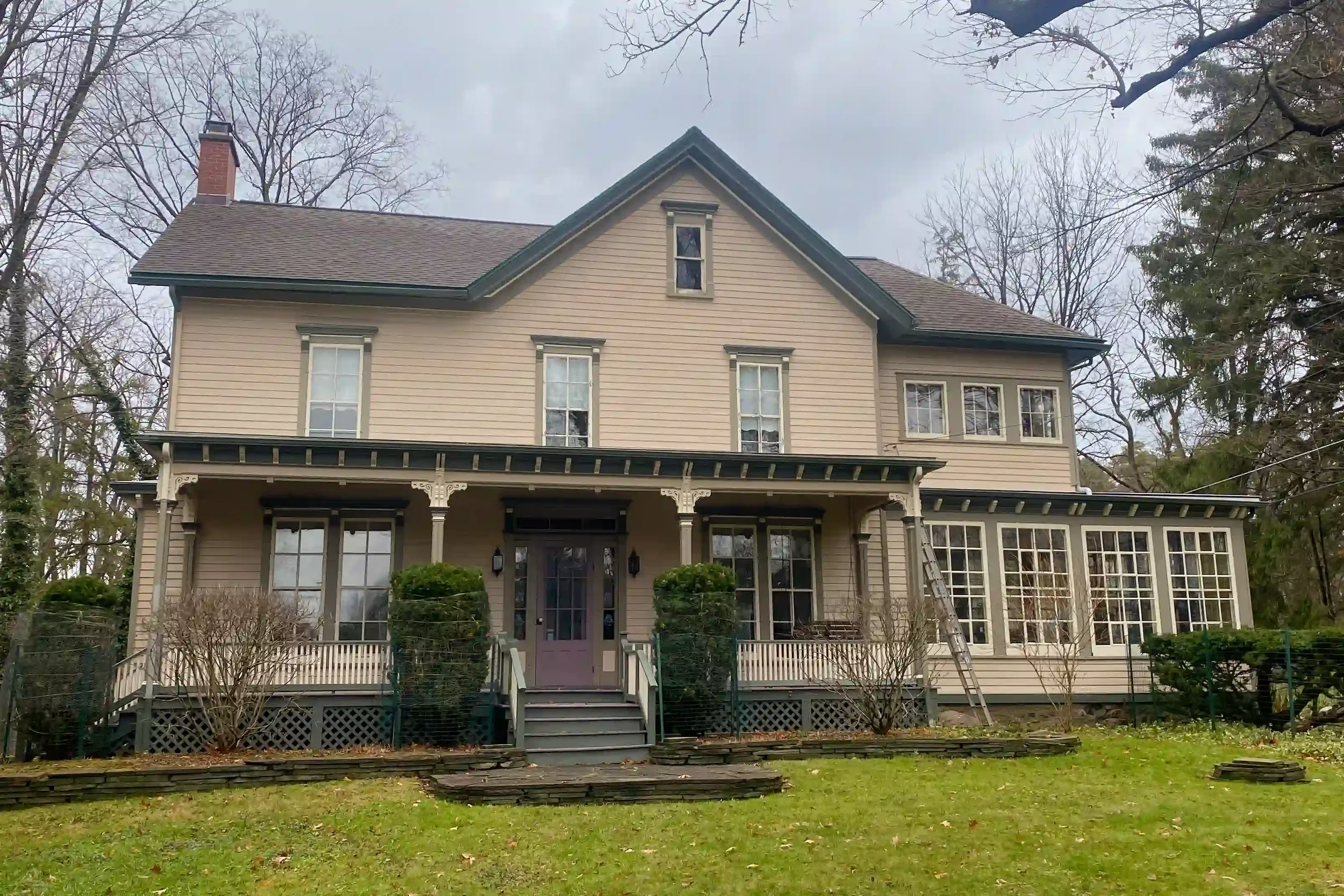 A large, beige, Victorian-style house with a gabled roof, multiple windows, and a porch with decorative trim. The yard has some small bushes, a lawn, leafless trees, and a cloudy sky.
