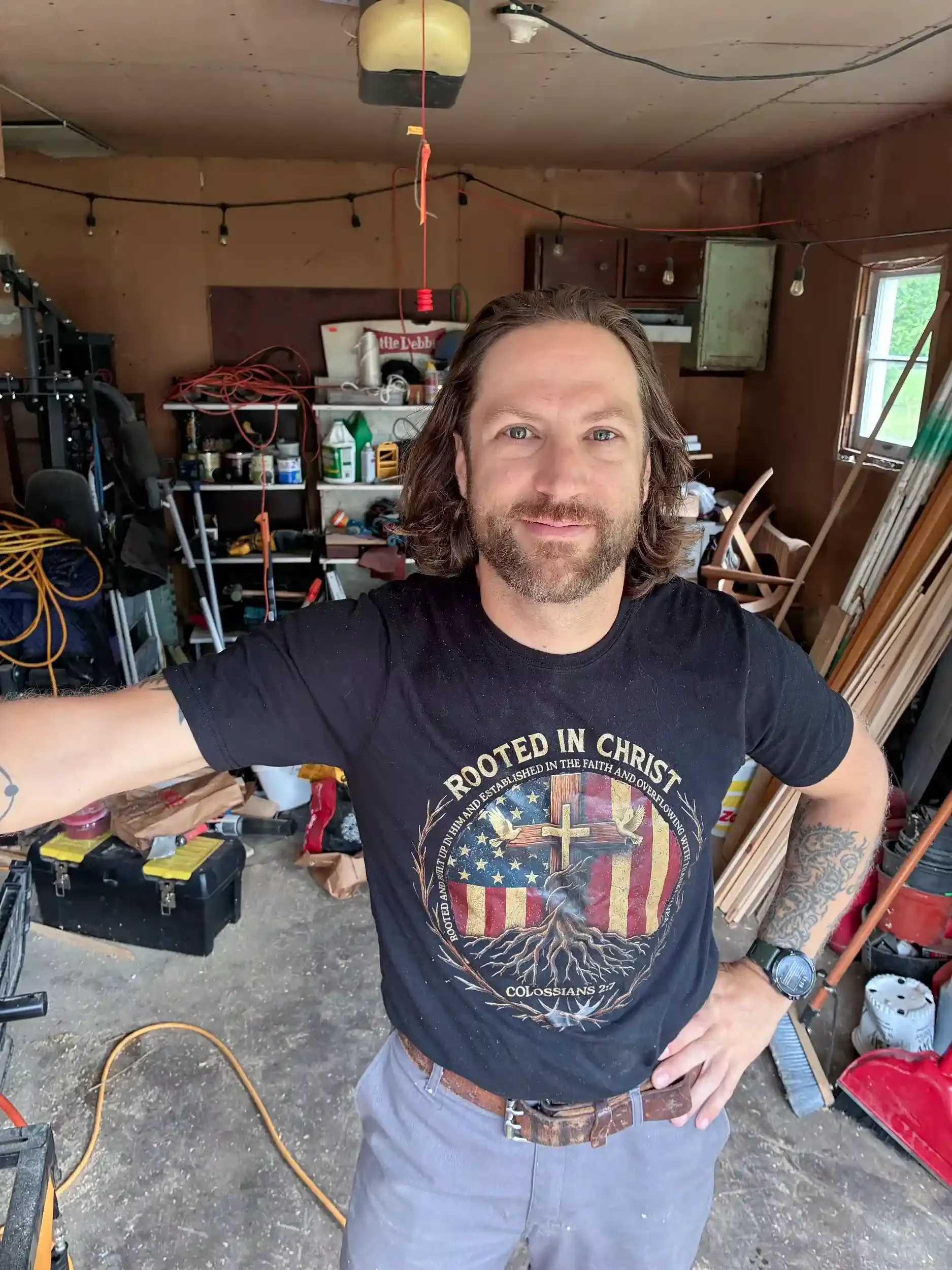 A man with long hair and a beard wearing a black T-shirt with religious and patriotic imagery, standing inside a cluttered garage. The garage contains various tools, cords, and hardware on shelves, with a window to the right allowing natural light. The man has a tattoo on his left forearm, wears a watch, and poses with his right hand on his hip and left arm extended.