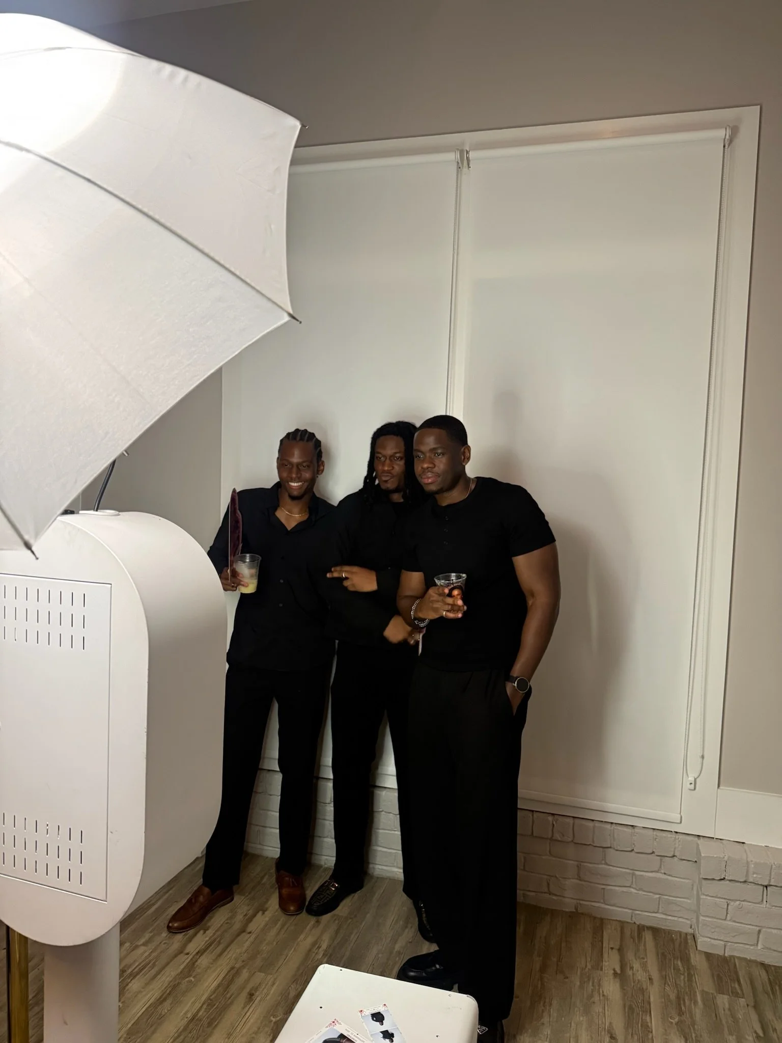Three men dressed in black standing together indoors, holding drinks, in front of a white wall with closed blinds and a brick baseboard.
