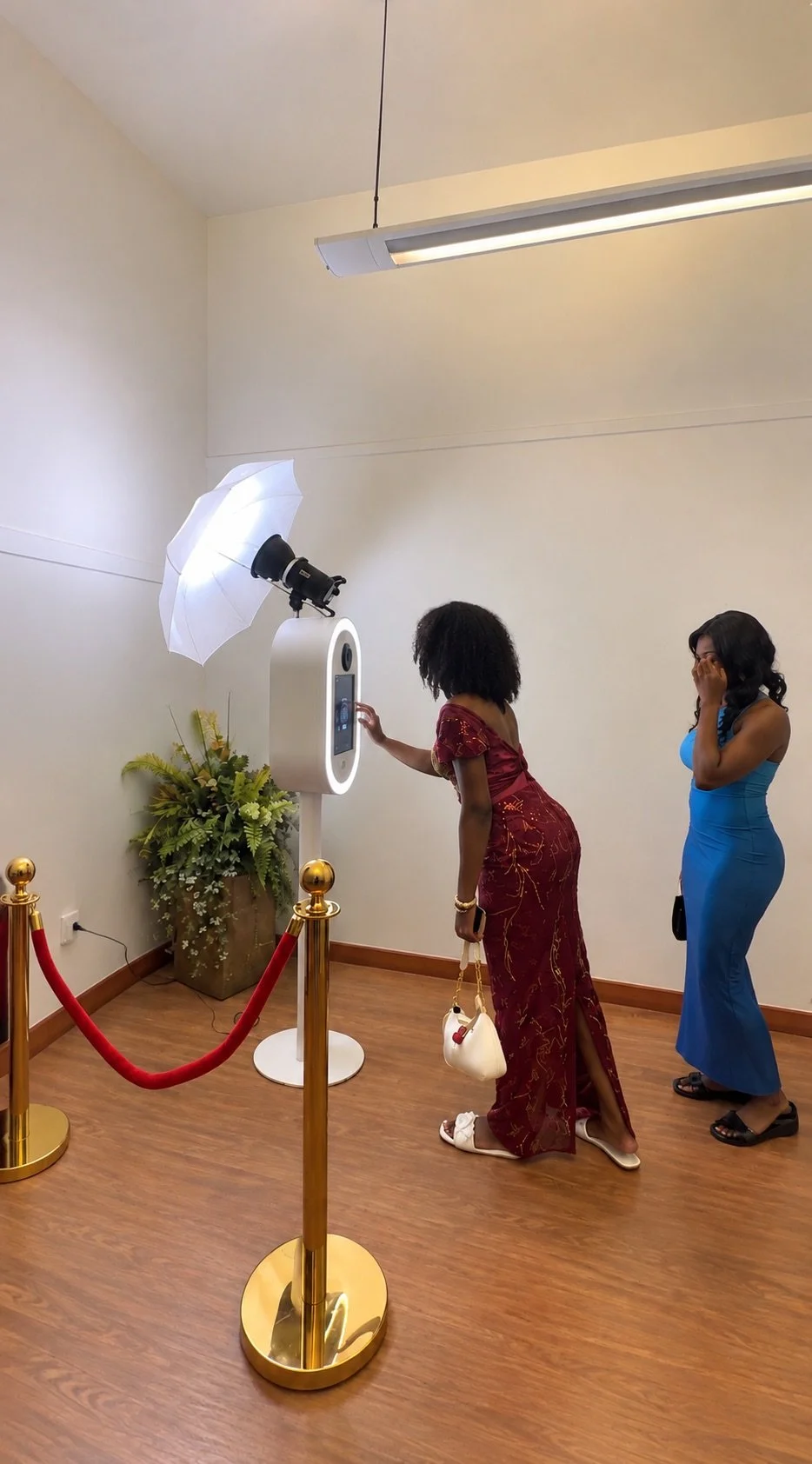 Two women standing inside a photo booth, one in a red dress adjusting the booth and the other in a blue dress covering her face, with photo studio equipment including a ring light and umbrella reflected in the booth.