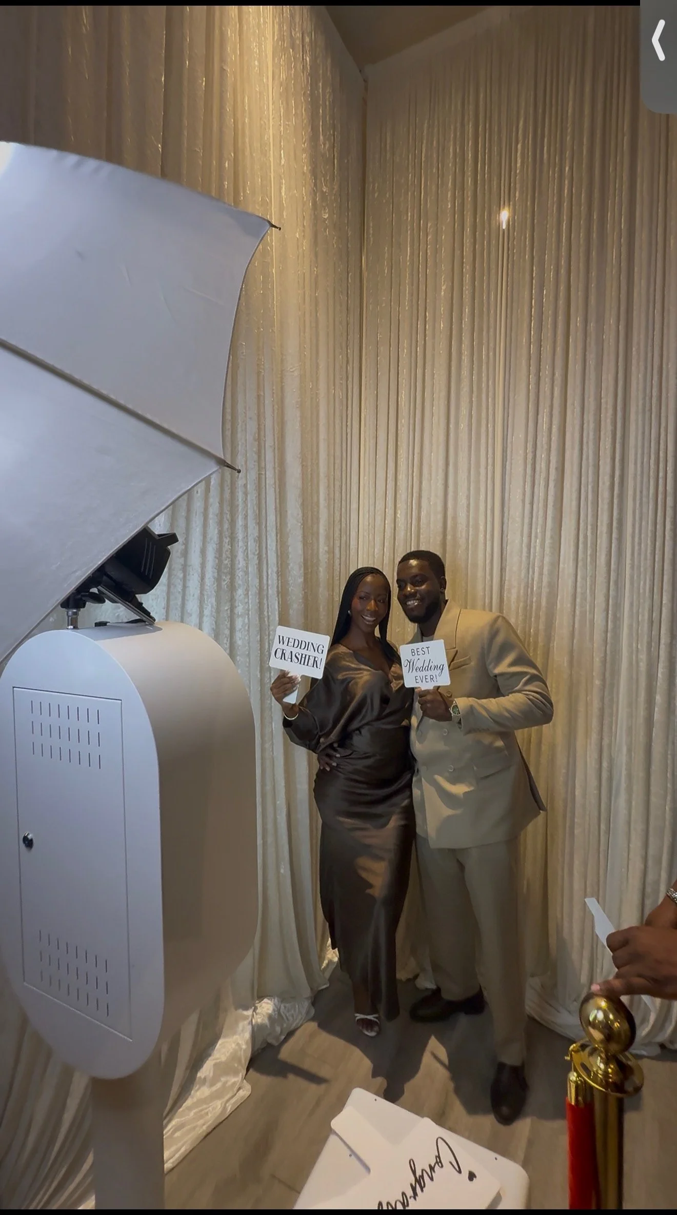 A joyful couple dressed in formal attire poses at a wedding reception, holding signs that read "WEDDING CASHIER!" and "BEST WEDDING EVER!". They stand in front of beige curtains, with a photo booth setup nearby, and a stanchion with a velvet rope visible in the foreground.