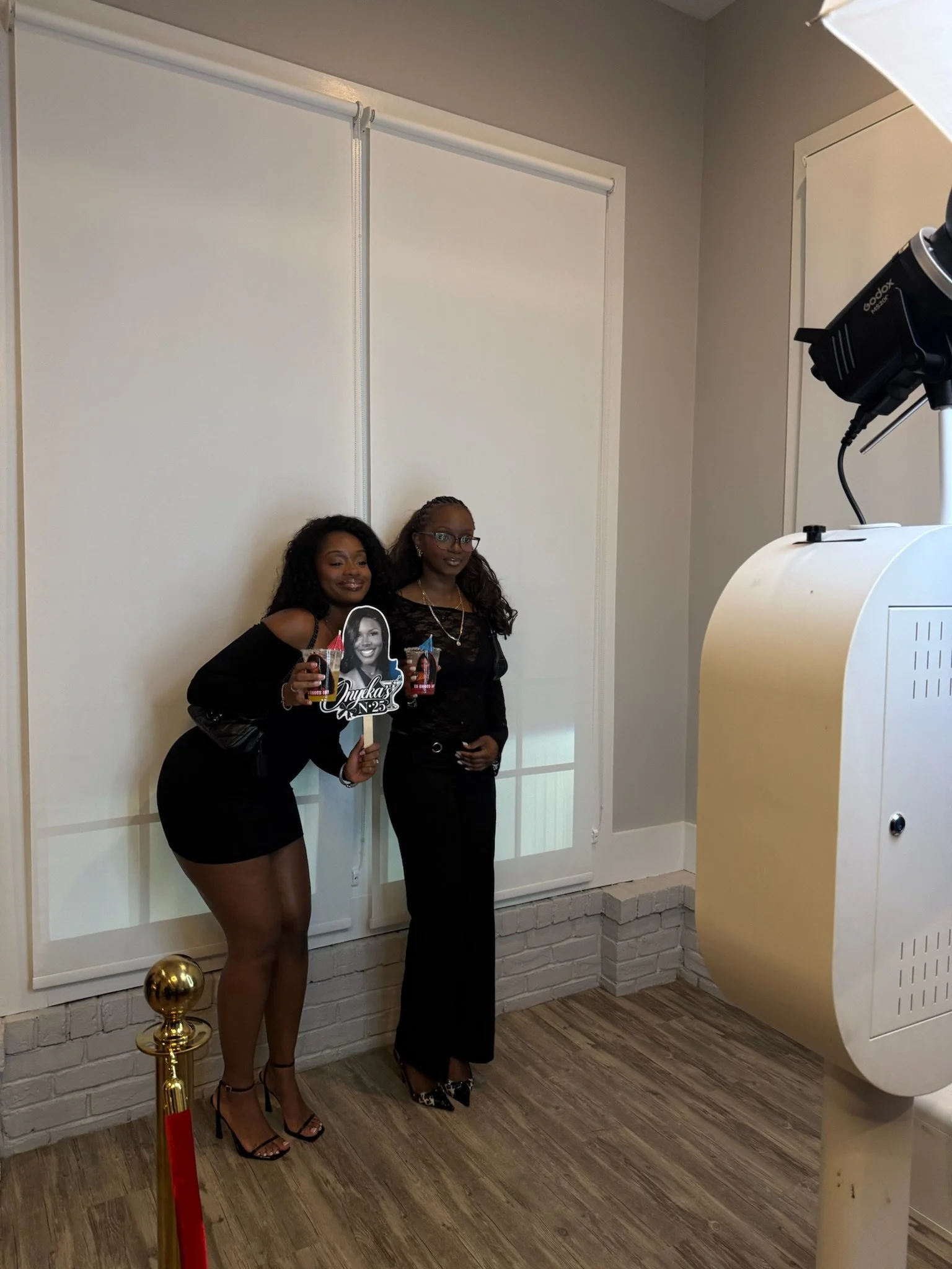 Two women dressed in black outfits pose for a photo in front of a white window cover, holding drinks and a framed black-and-white photo. One woman has glasses and braided hair, both are smiling. To the right, part of a large coffee roasting machine is visible.