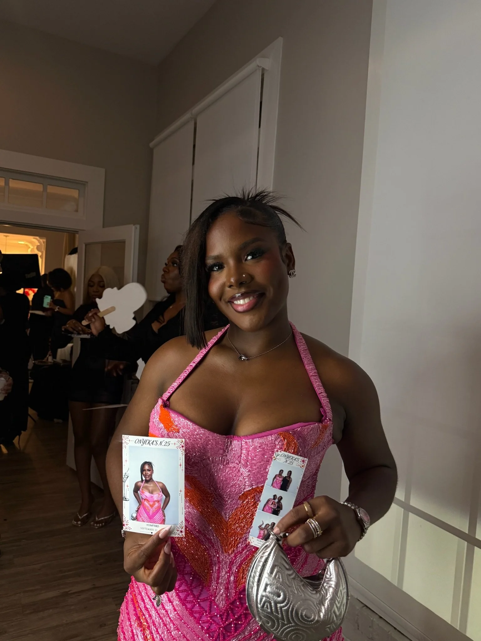 A woman with dark hair smiling at a celebration, holding a photo strip of herself in a pink dress, with other women in black outfits in the background, inside a decorated room.