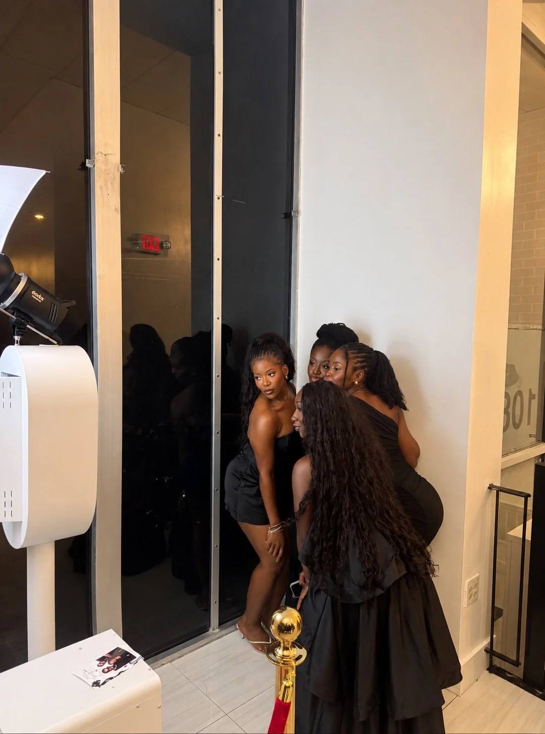 Four women dressed in black outfits posing for a photo near a large mirror, with a photographer capturing the picture. They are standing by a white wall and glass, with a small table and a poster nearby.