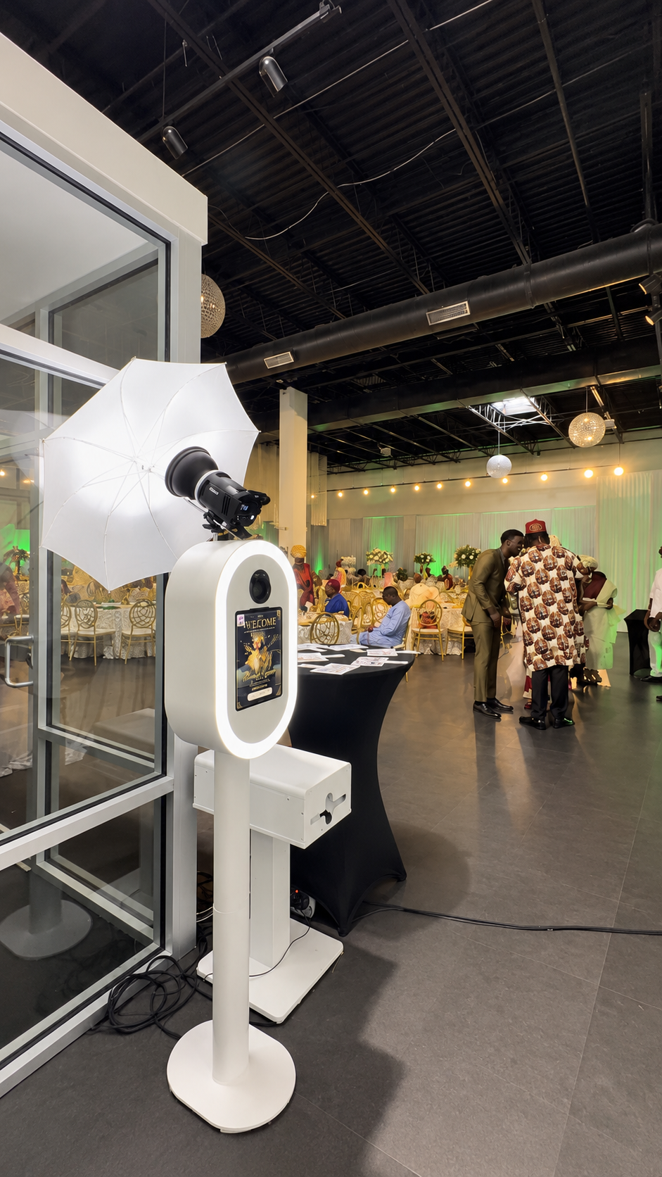 Photo of a photo booth with a white umbrella, camera, and digital screen at an indoor event with people socializing and dining in the background.