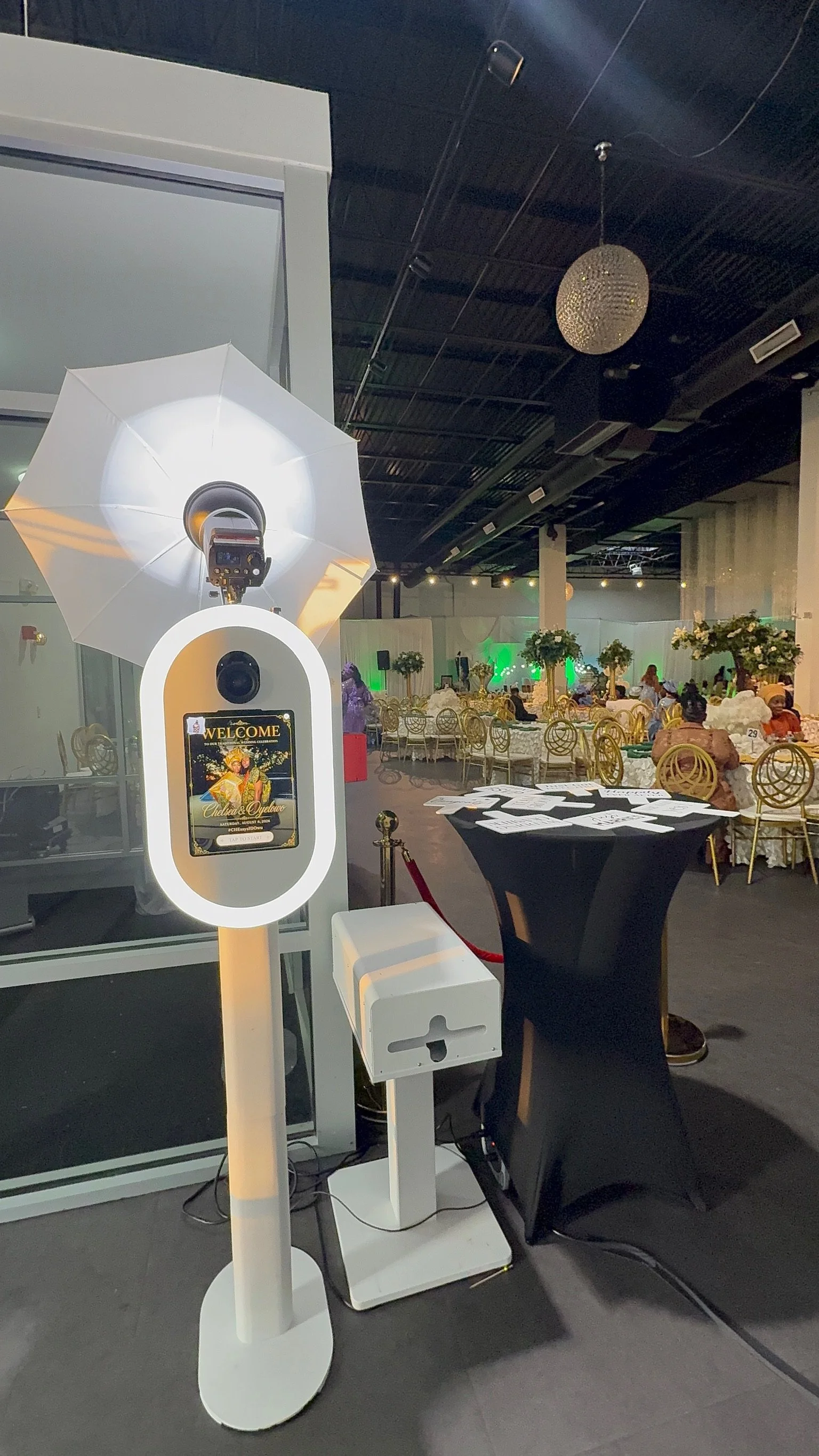 Photo of a photobooth with a white umbrella, digital camera, and a welcome sign at a decorated event space with tables, chairs, and floral centerpieces.