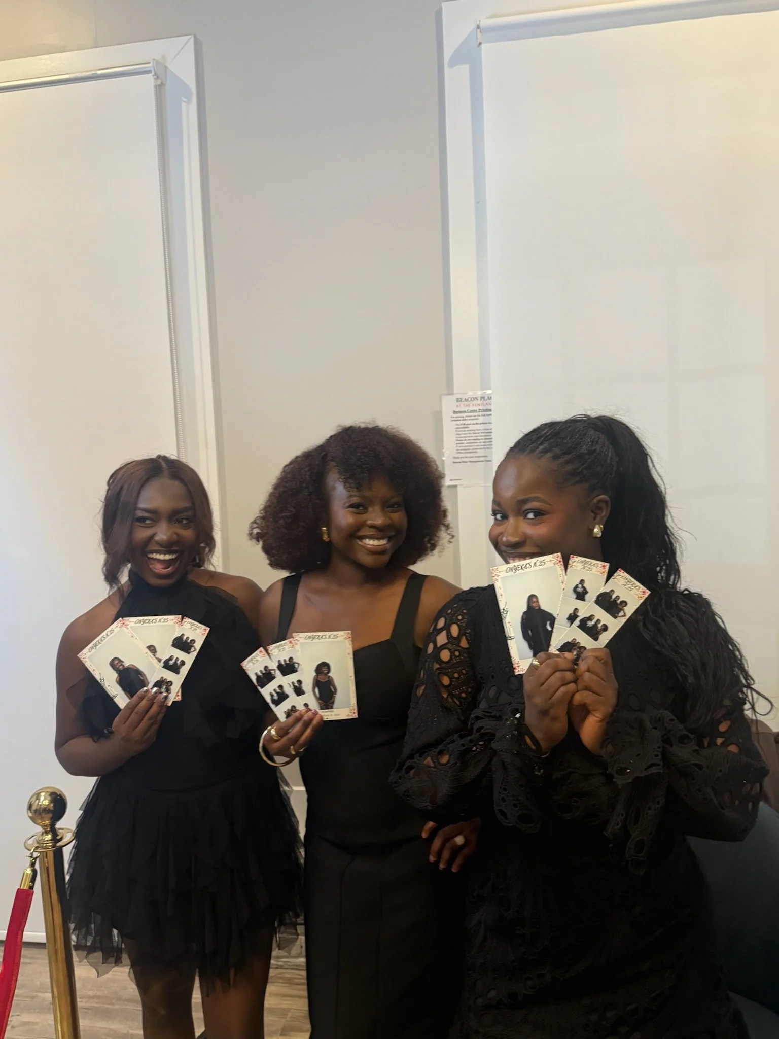 Three women in black dresses smiling and holding photo booth pictures, standing on a red carpet with a white wall background.