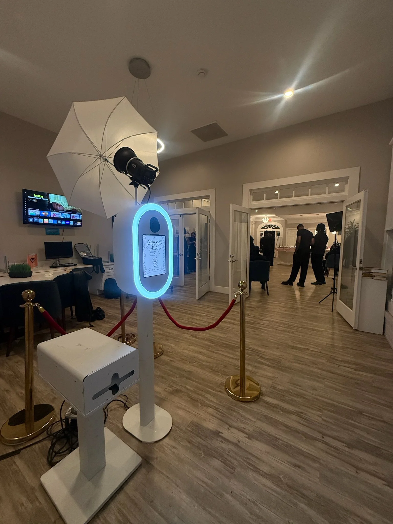 Photo of a photo booth setup with a white umbrella light on a stand, a sign with a blue neon outline, and a backdrop with people inside a room with open doors, wood flooring, and a wall-mounted television.