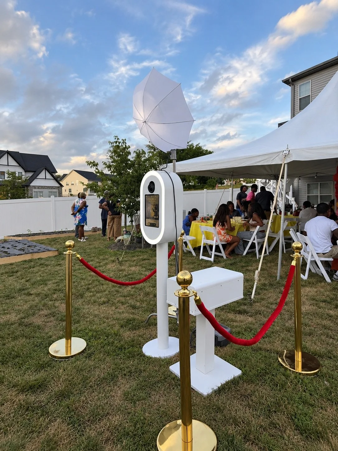 Outdoor gathering with a party tent, guests sitting at tables with yellow tablecloths, a photo booth station with a white umbrella, and a white fence in the background under a partly cloudy sky.