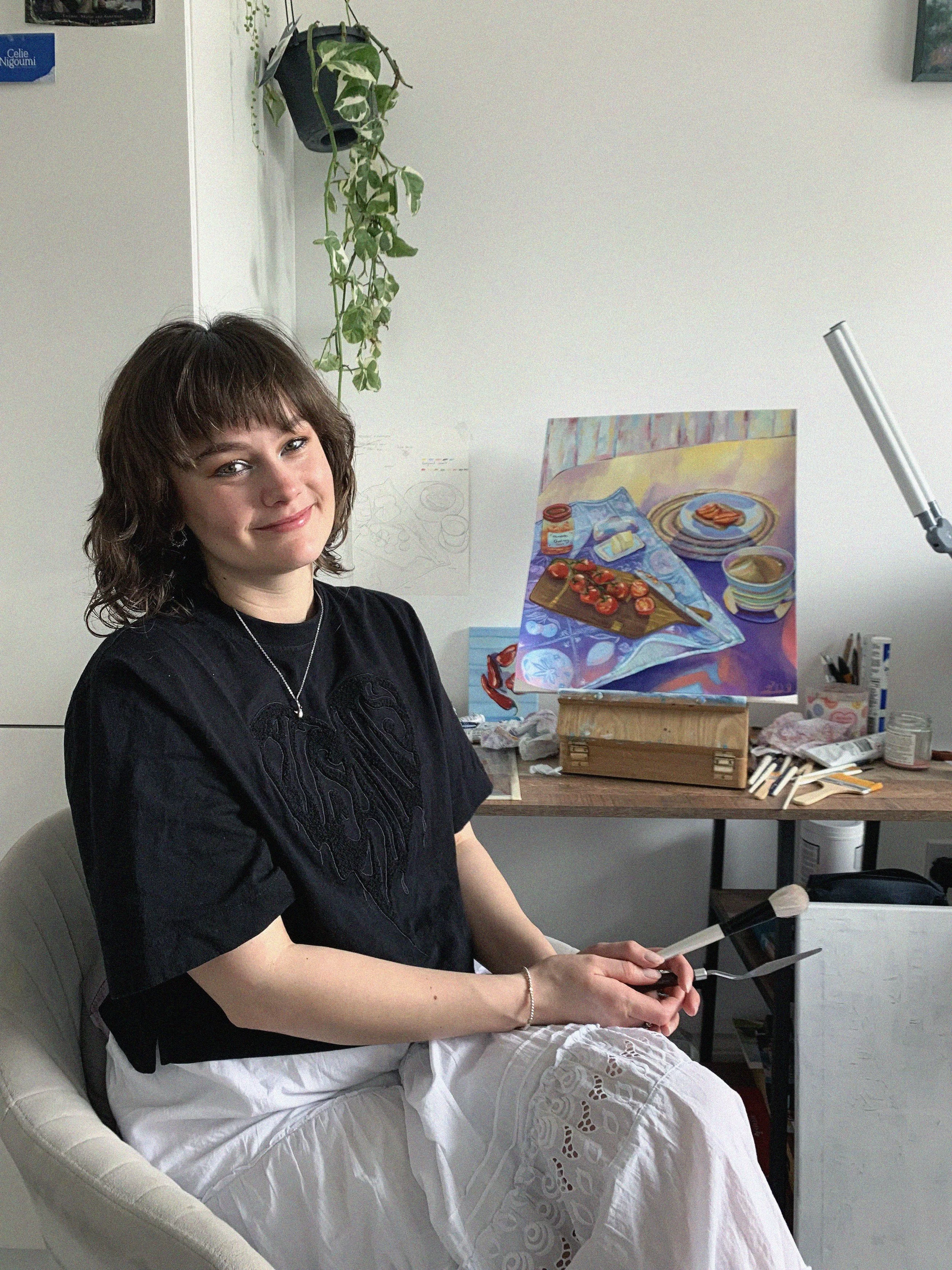 Young woman in a black T-shirt sitting on a bed and holding a tablet stylus, with a hanging variegated plant, artwork, and shelving in the background.