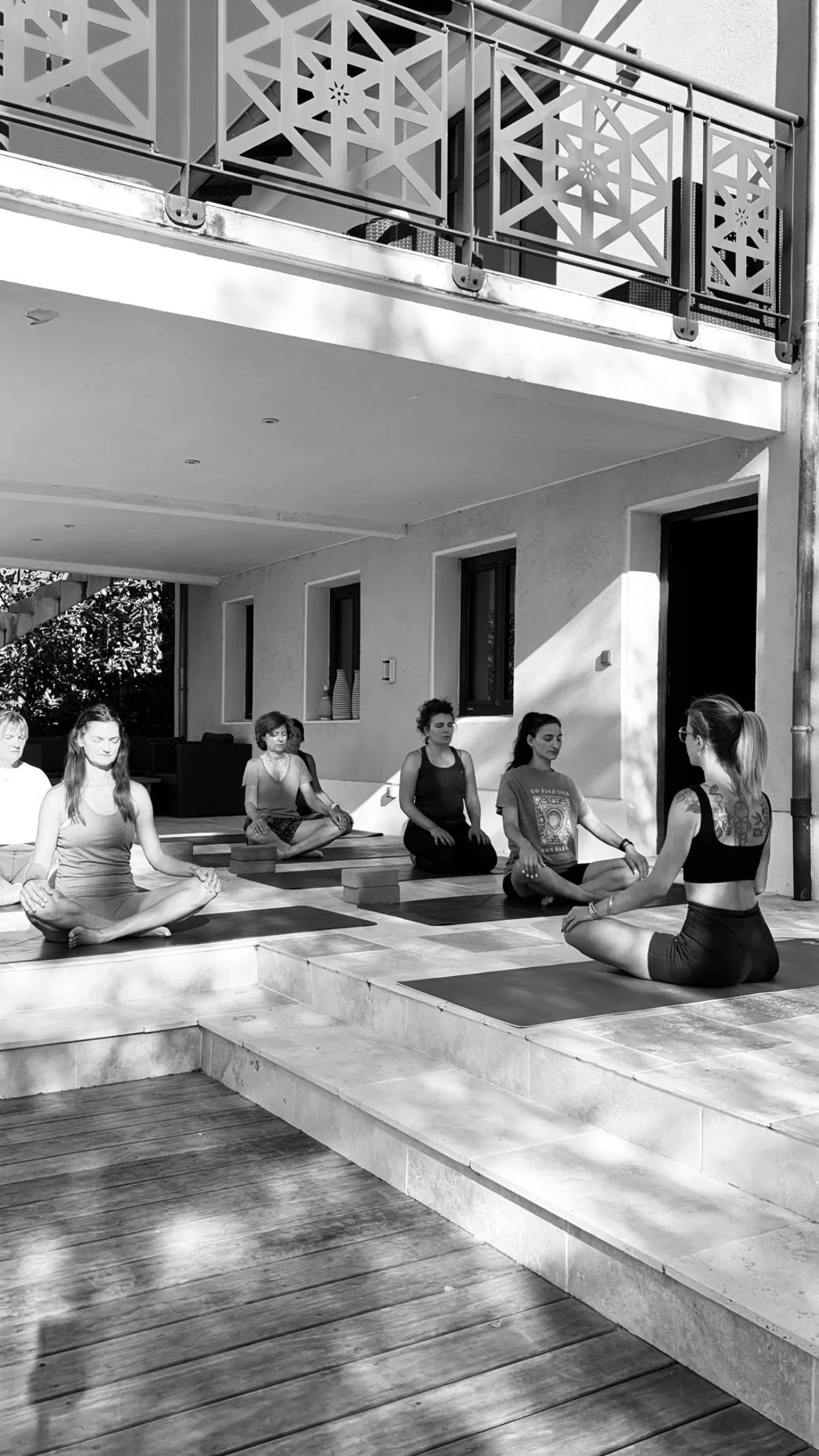 Groupe de personnes pratiquant le yoga en extérieur sur un podium en gravier, devant une maison avec un balcon en métal ajouré, en noir et blanc.