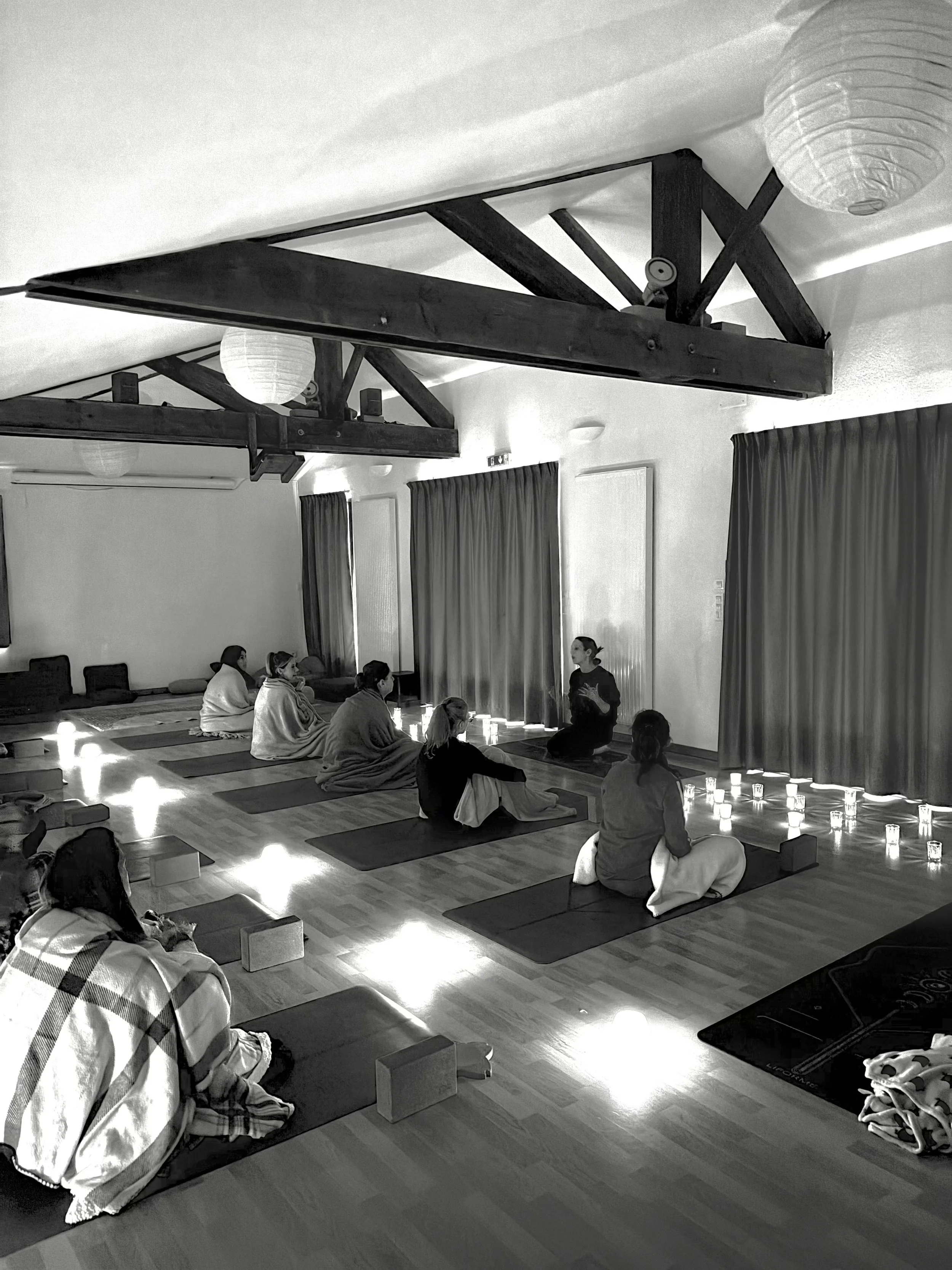 Une séance de yoga ou de méditation en groupe dans une salle avec des chandelles, des tapis de yoga, des coussins et des lanternes suspendues au plafond. Une instructrice explique aux participants.