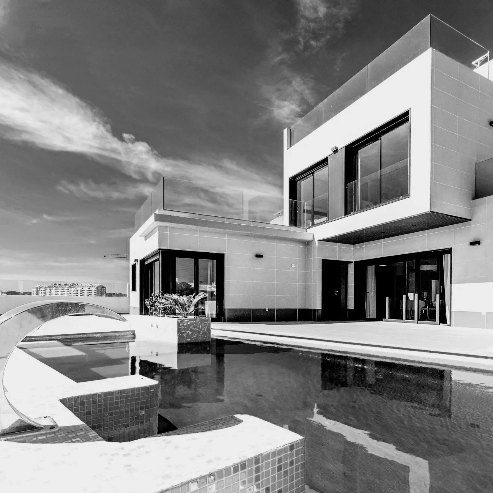 Une vue d'une maison moderne avec une piscine en premier plan, un bâtiment blanc avec grandes fenêtres derrière, et un ciel nuageux en arrière-plan.