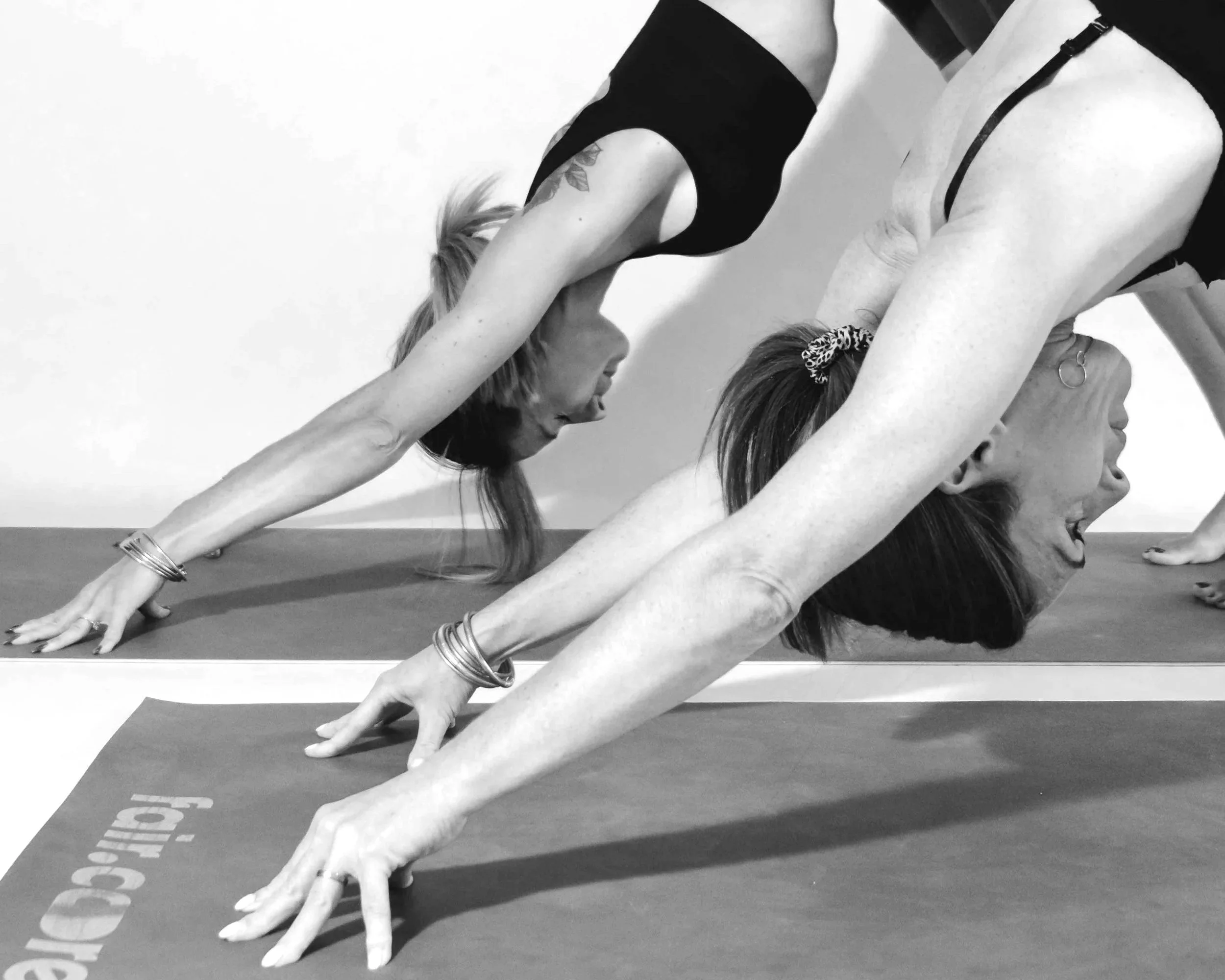Deux femmes font du yoga en position invertée sur un tapis, l'une au-dessus de l'autre, dans une salle blanche.