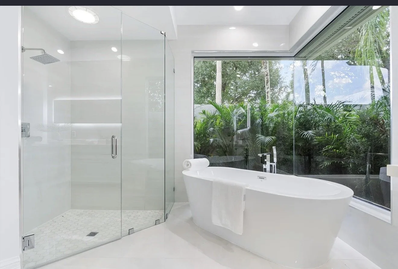 Modern bathroom with a glass-enclosed shower and a soaking bathtub by a large window with a view of lush green plants.