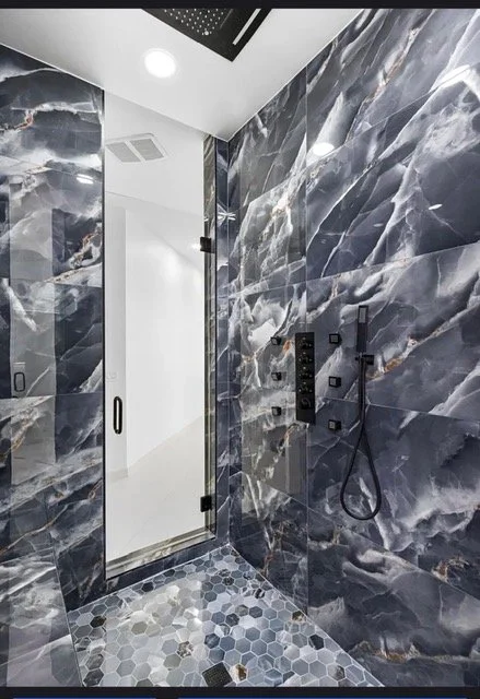 Modern walk-in shower with blue and gray marble walls, hexagonal gray floor tiles, black shower fixtures, and a glass door.