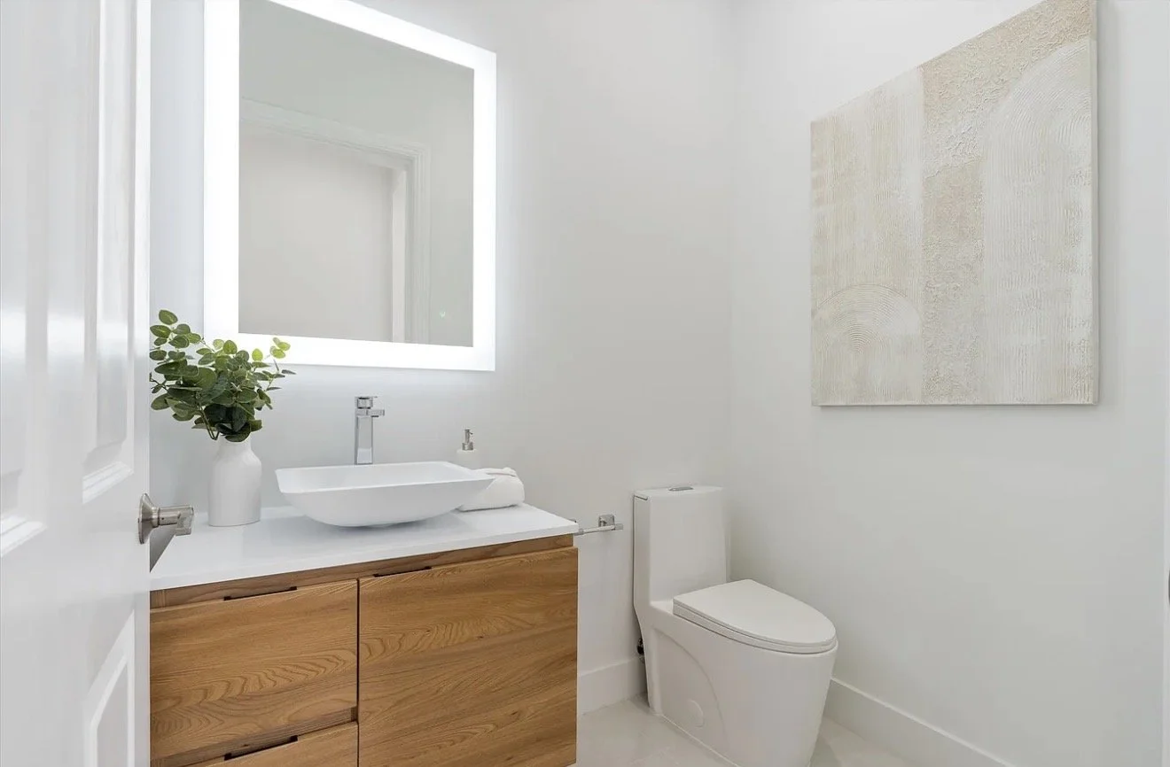 Modern bathroom with white walls, a wooden vanity with a white countertop, a vessel sink, a large backlit mirror, a potted green plant, a white toilet, and a textured abstract art piece on the wall.