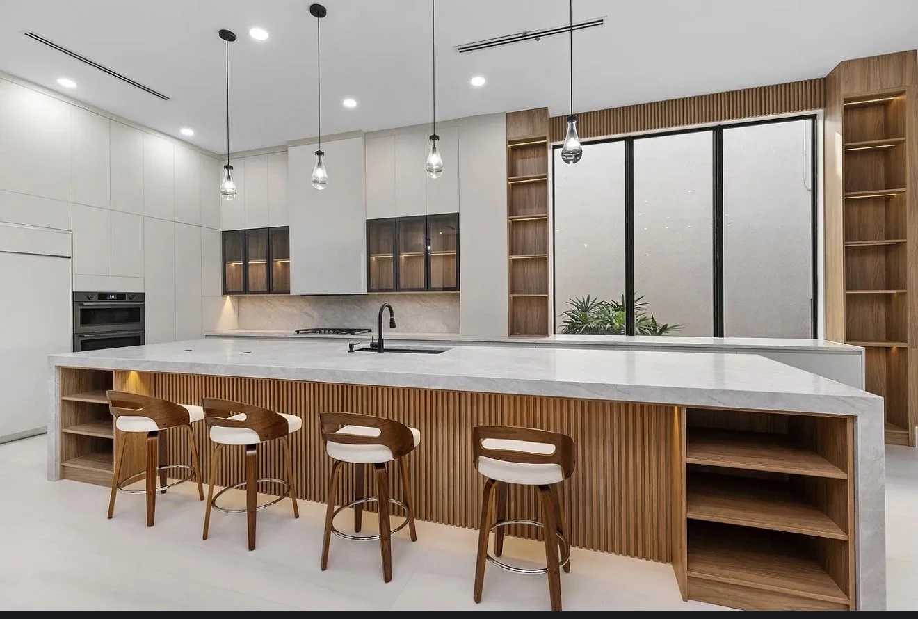 Modern kitchen with white cabinetry, wood accents, marble island, four bar stools with wood and white cushions, pendant lights, and large window with plants outside.