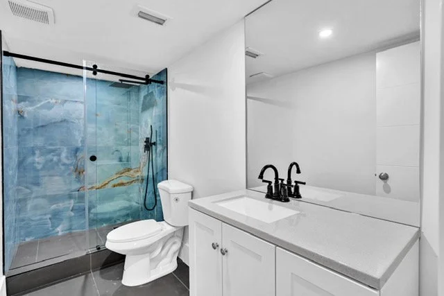Bathroom with a glass-enclosed shower, white toilet, white vanity with a sink, black fixtures, and a large mirror.