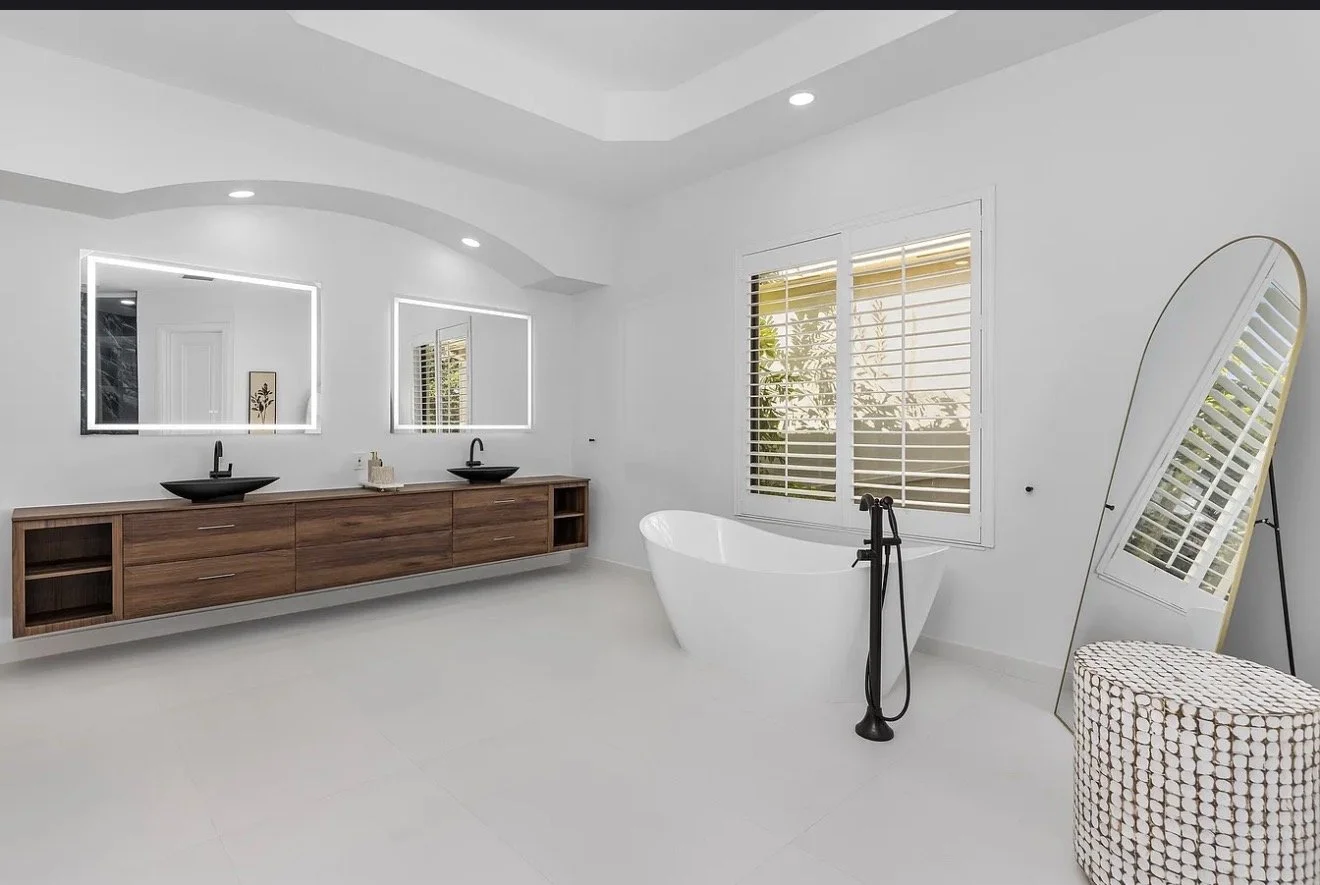 Modern bathroom with white walls and floor, double vanity with black vessel sinks, large mirror with LED lighting, freestanding bathtub, window with blinds, and a full-length mirror leaning against the wall.