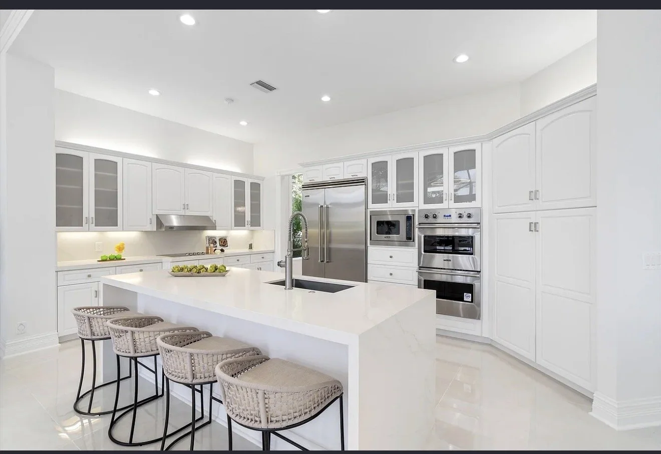 Modern white kitchen with a waterfall island, beige barstools, stainless steel appliances, and bright lighting.