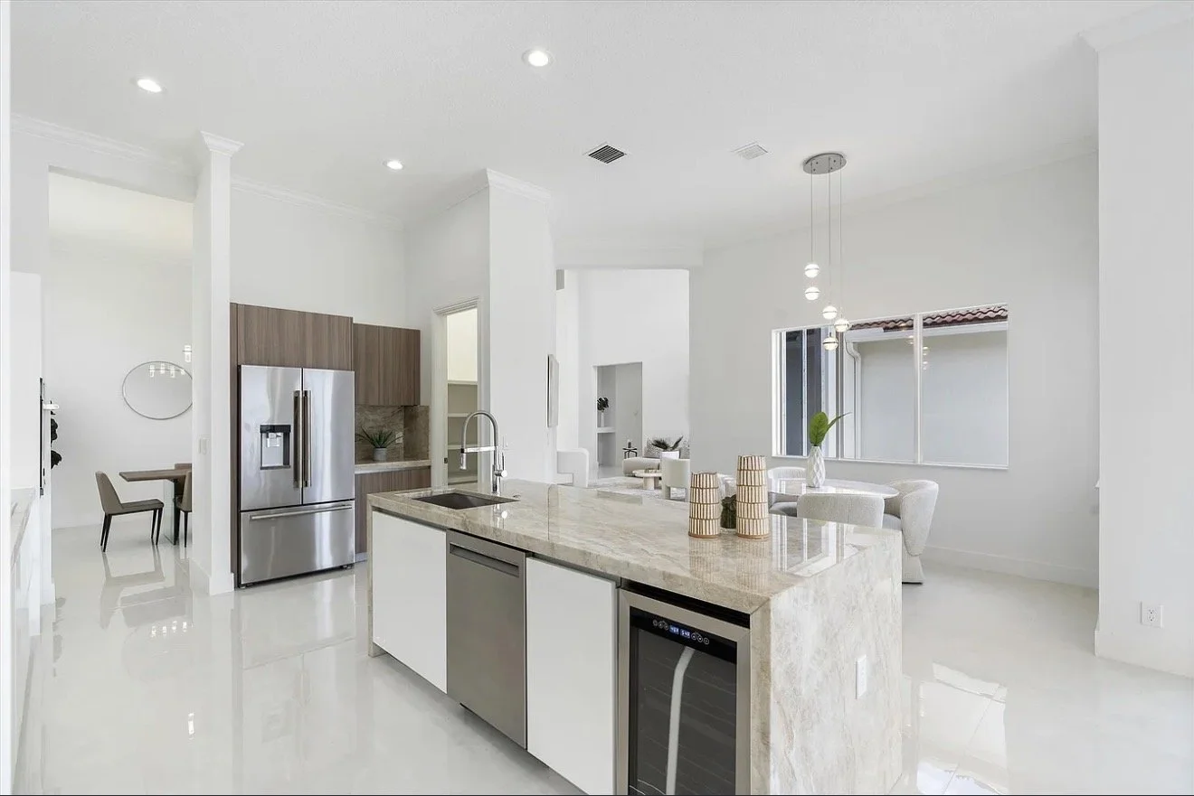 Modern, minimalist kitchen with white walls, a marble island, stainless steel refrigerator, and a dining area with white chairs and a hanging light fixture.