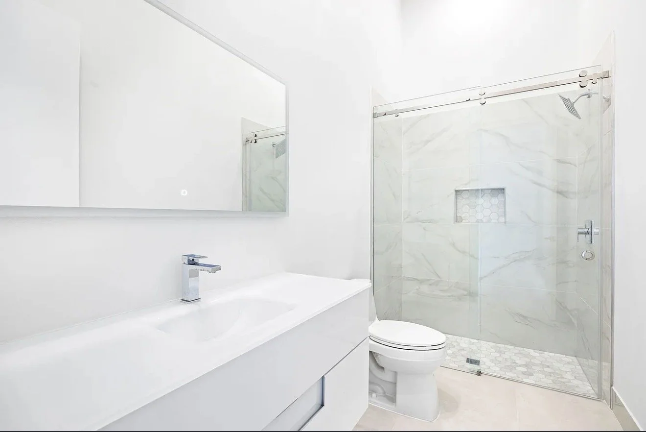 Minimalist white bathroom with a large wall mirror, a white vanity with a sleek faucet, a white toilet, and a glass shower with marble tiles and a hexagonal tile shower floor.