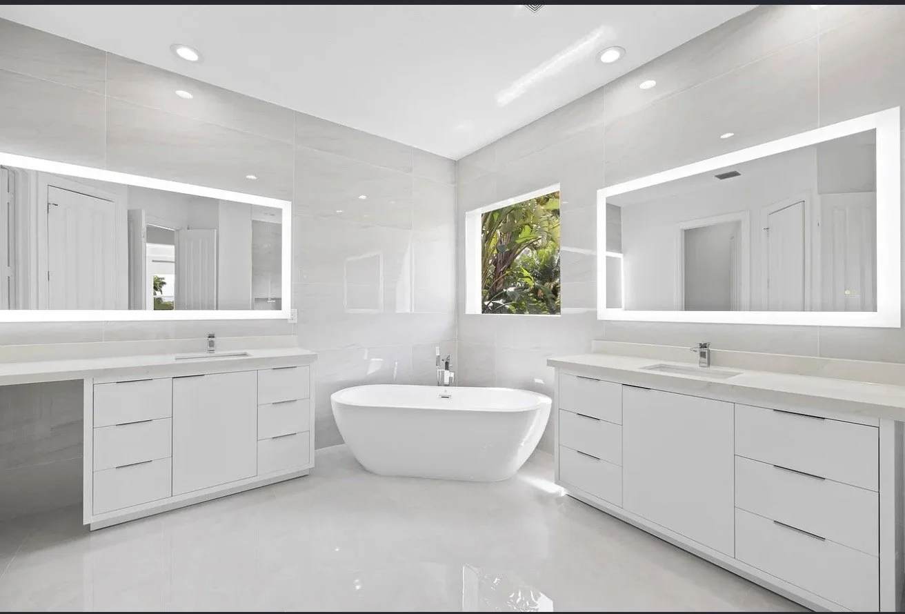 Modern white bathroom with double vanities, large mirrors, a freestanding bathtub, and a window with greenery outside.