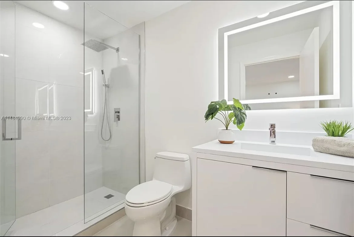 Modern white bathroom featuring a glass-enclosed shower, a white toilet, a white vanity with a large backlit mirror, and green plants on the vanity countertop.