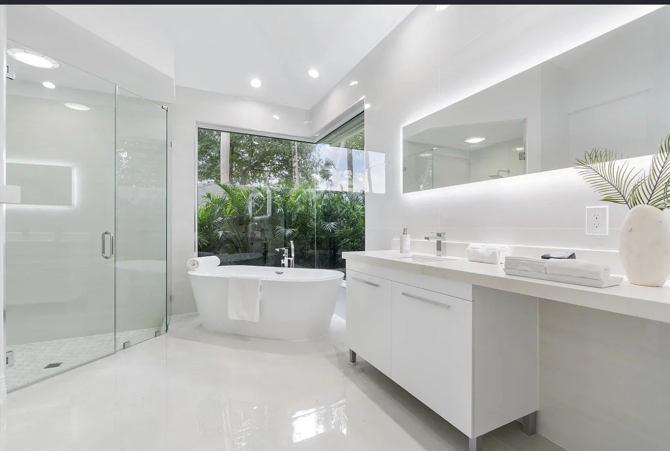 Modern white bathroom featuring a glass-enclosed shower, a free-standing bathtub with a view of greenery outside, a large mirror with LED lighting, a white vanity with towels and toiletries, and a potted plant for decoration.