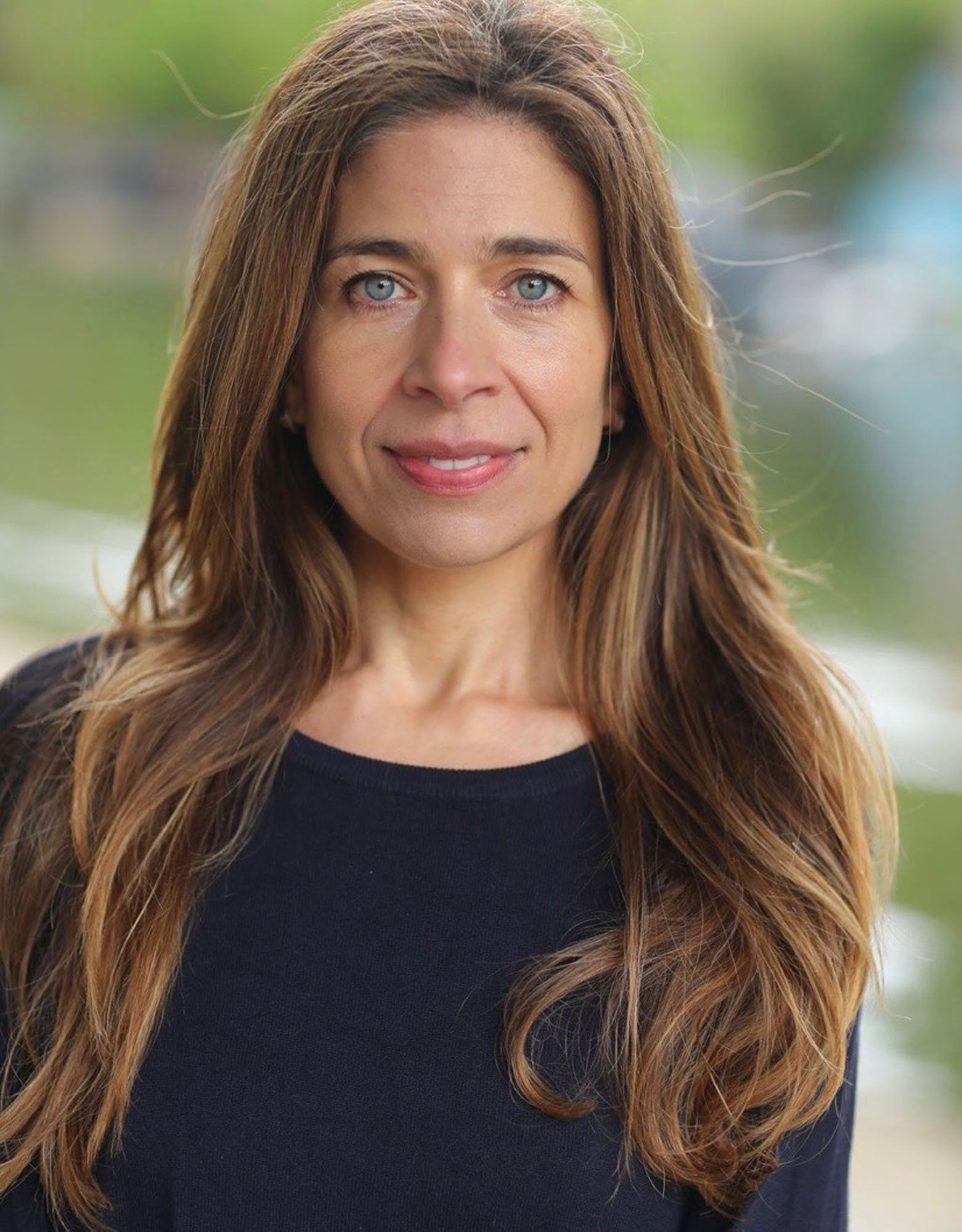 Close-up photo of a woman with long, wavy brown hair with blonde highlights, light blue eyes, and a slight smile, outdoors with blurred green and blue background.
