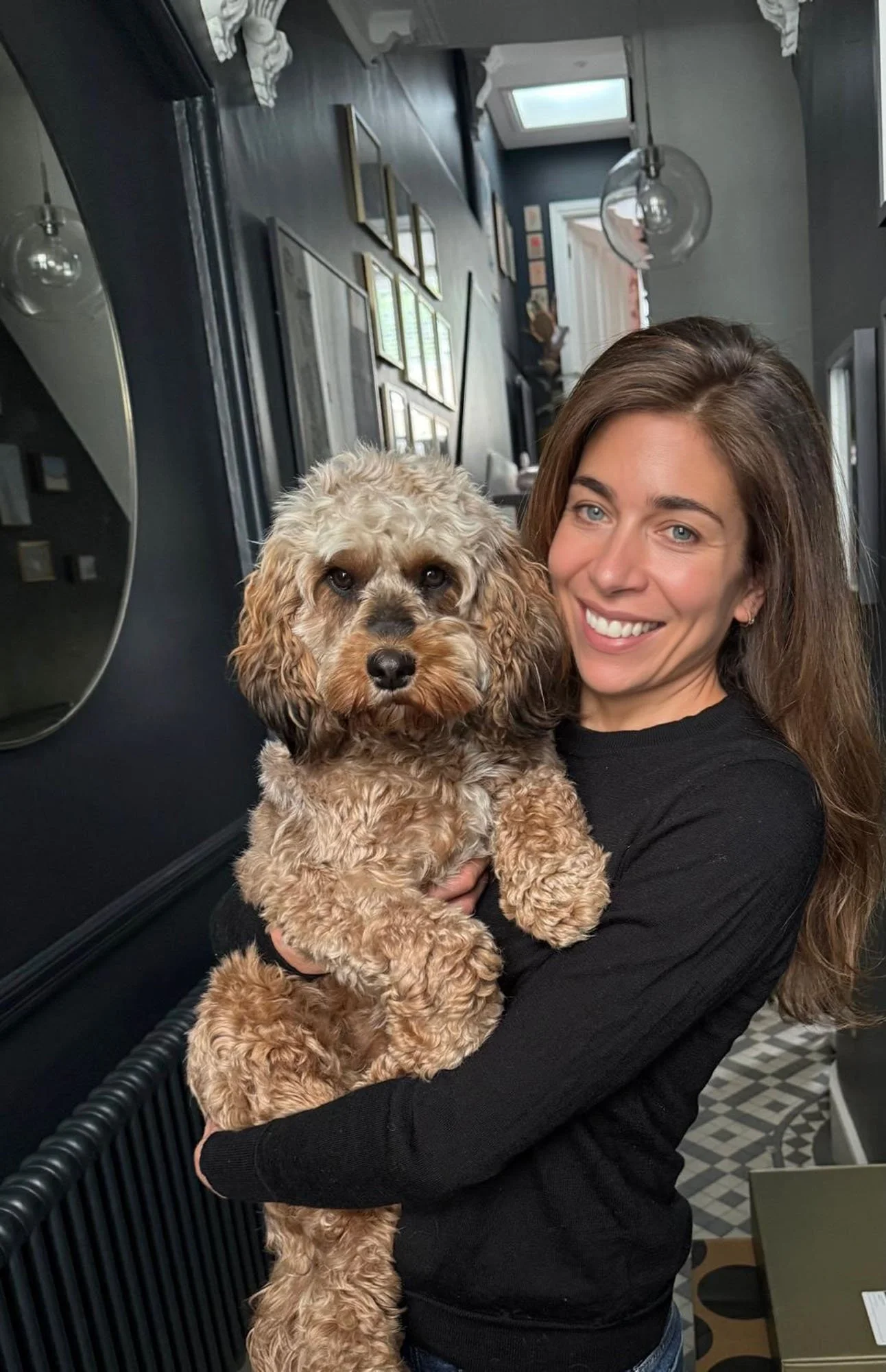 A woman is holding a large, curly-haired dog while smiling at the camera inside a stylish room with dark walls and decorative lighting.