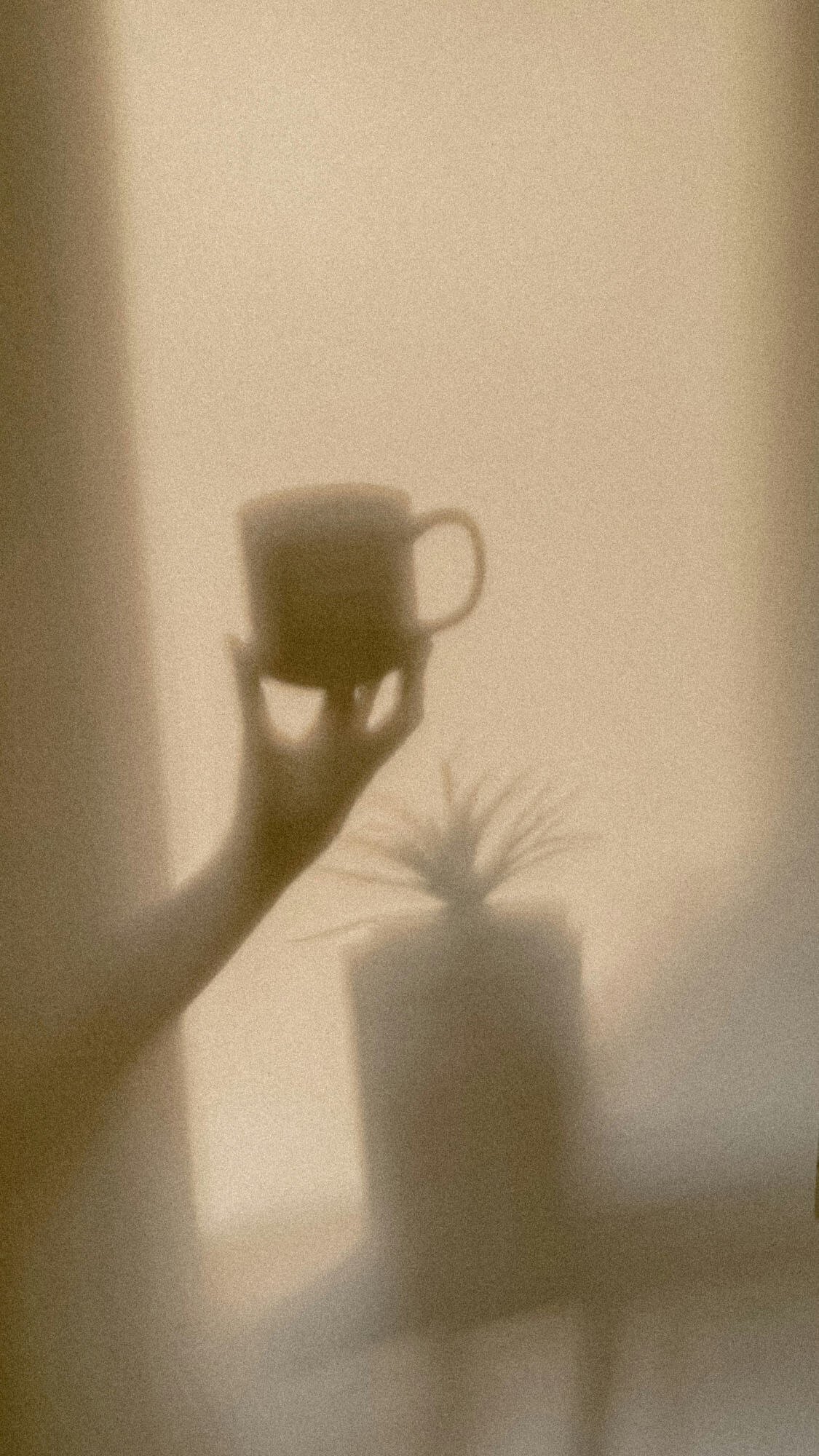 Shadow of a hand holding a coffee mug cast on a wall, with part of a person's foot and pants visible at the bottom.