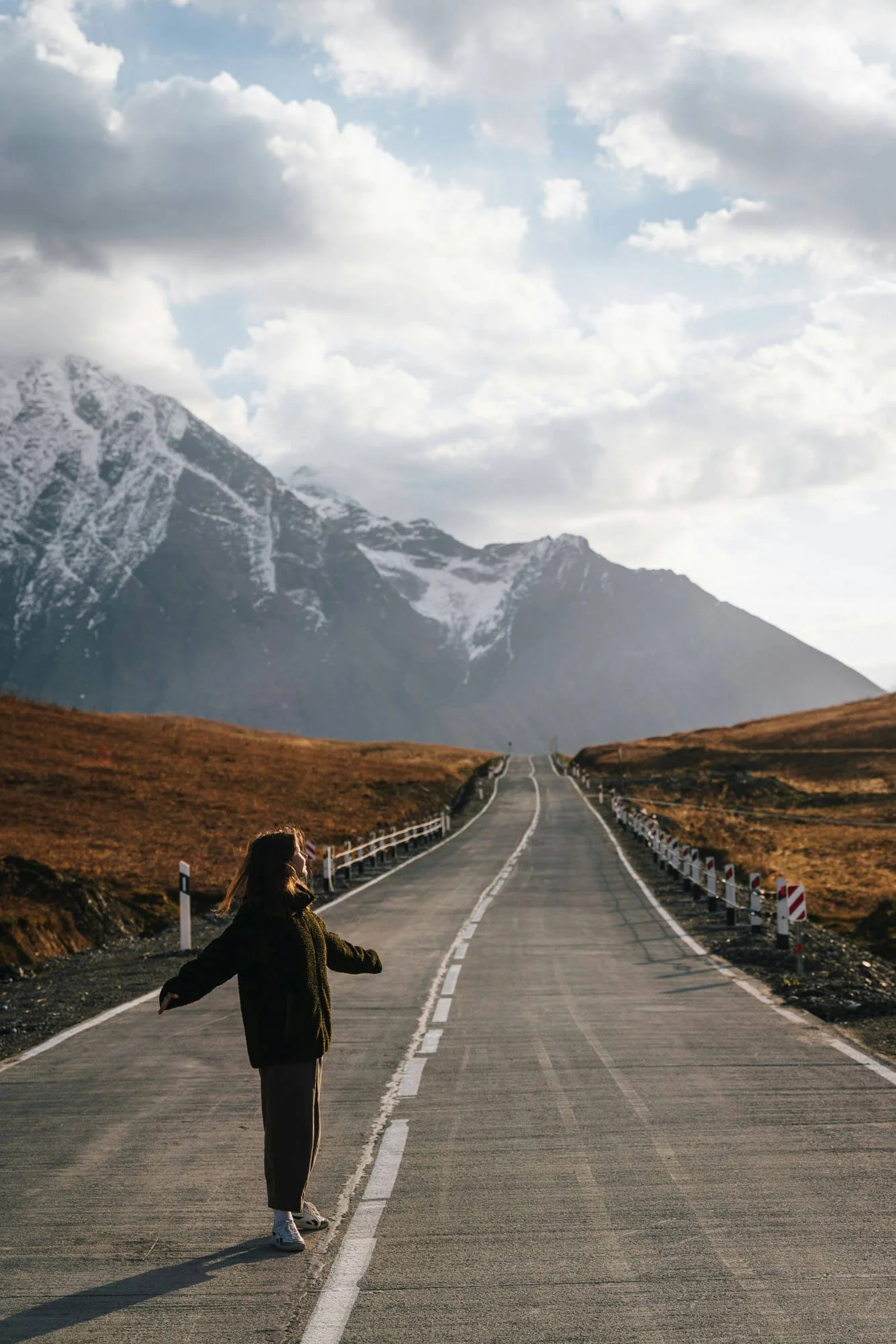 A person with long hair, wearing a dark coat and white sneakers, standing in the middle of a long, empty mountain road with snow-capped mountains in the background under cloudy sky.