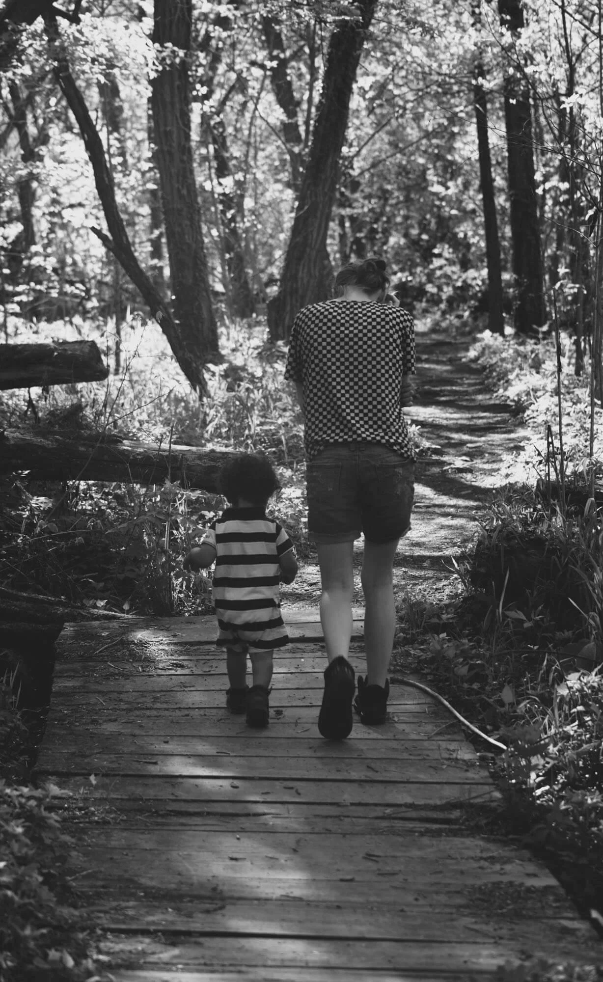 A woman and a young child walk together along a wooden pathway in a forest, surrounded by trees and foliage.
