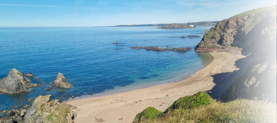 Coastal scene with sandy beach, rocky shoreline, blue ocean, and green cliffs under a clear sky.