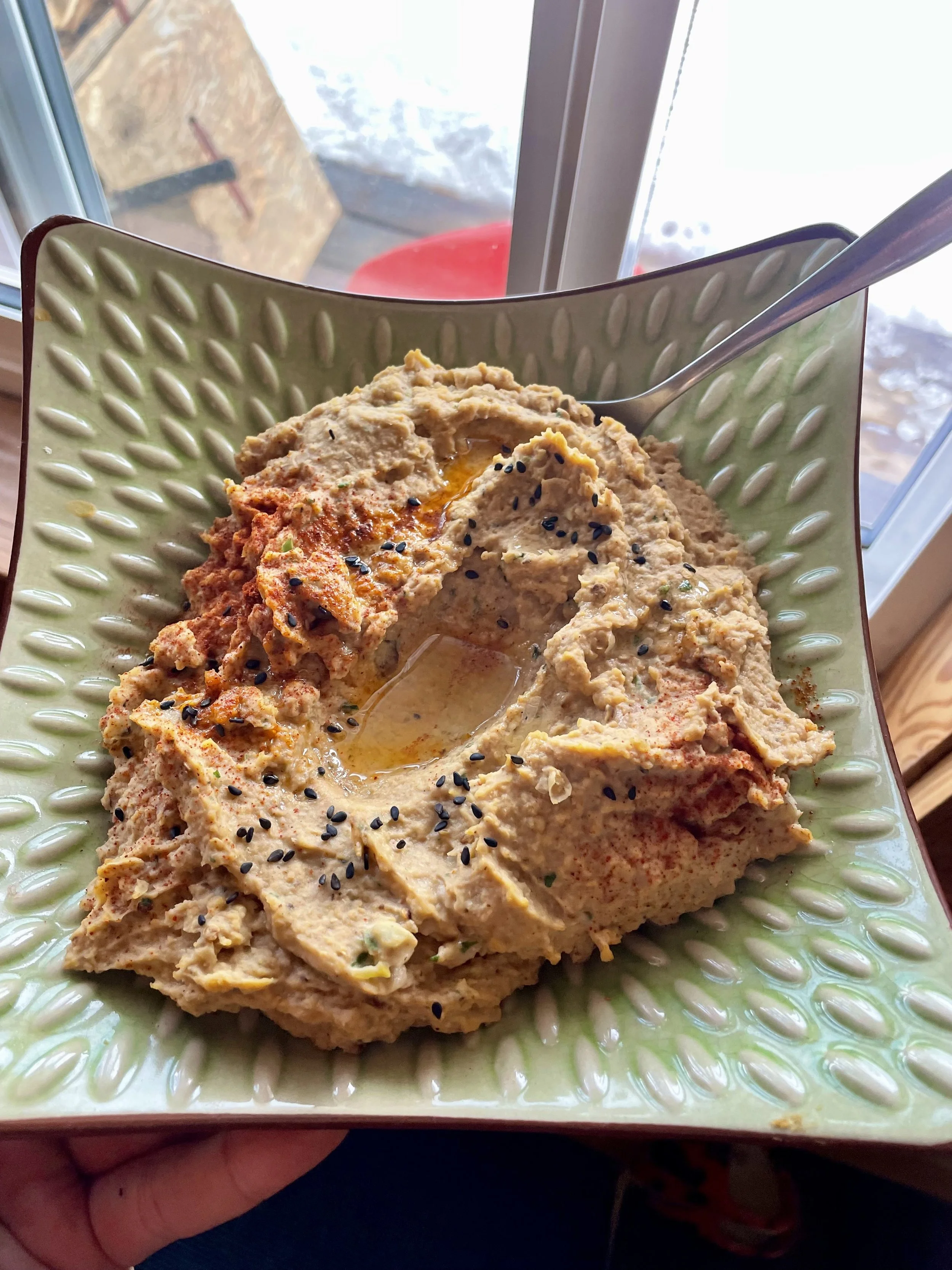 A bowl of hummus with black sesame seeds on top, placed on a decorative green plate, with a spoon inside the bowl. The background shows a window with sunlight prepared by a Revelstoke private chef.