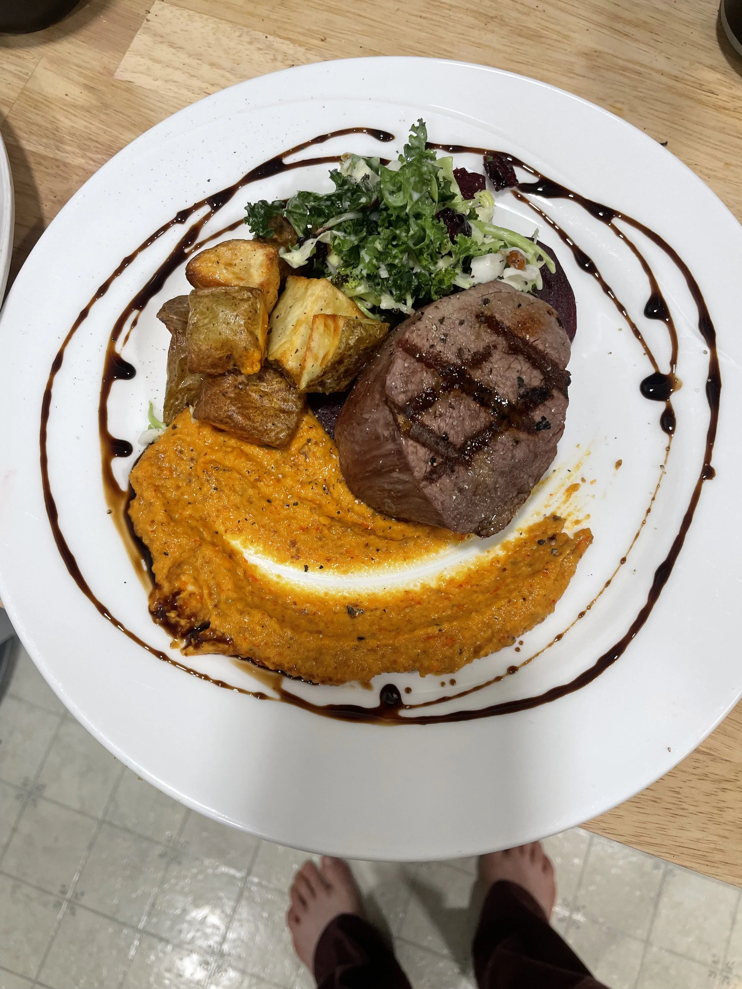A white plate of food containing grilled meat, roasted potatoes, a portion of mashed pumpkin, and a salad with leafy greens. The plate is decorated with a drizzle of balsamic glaze around the edges prepared by a Revelstoke private chef.