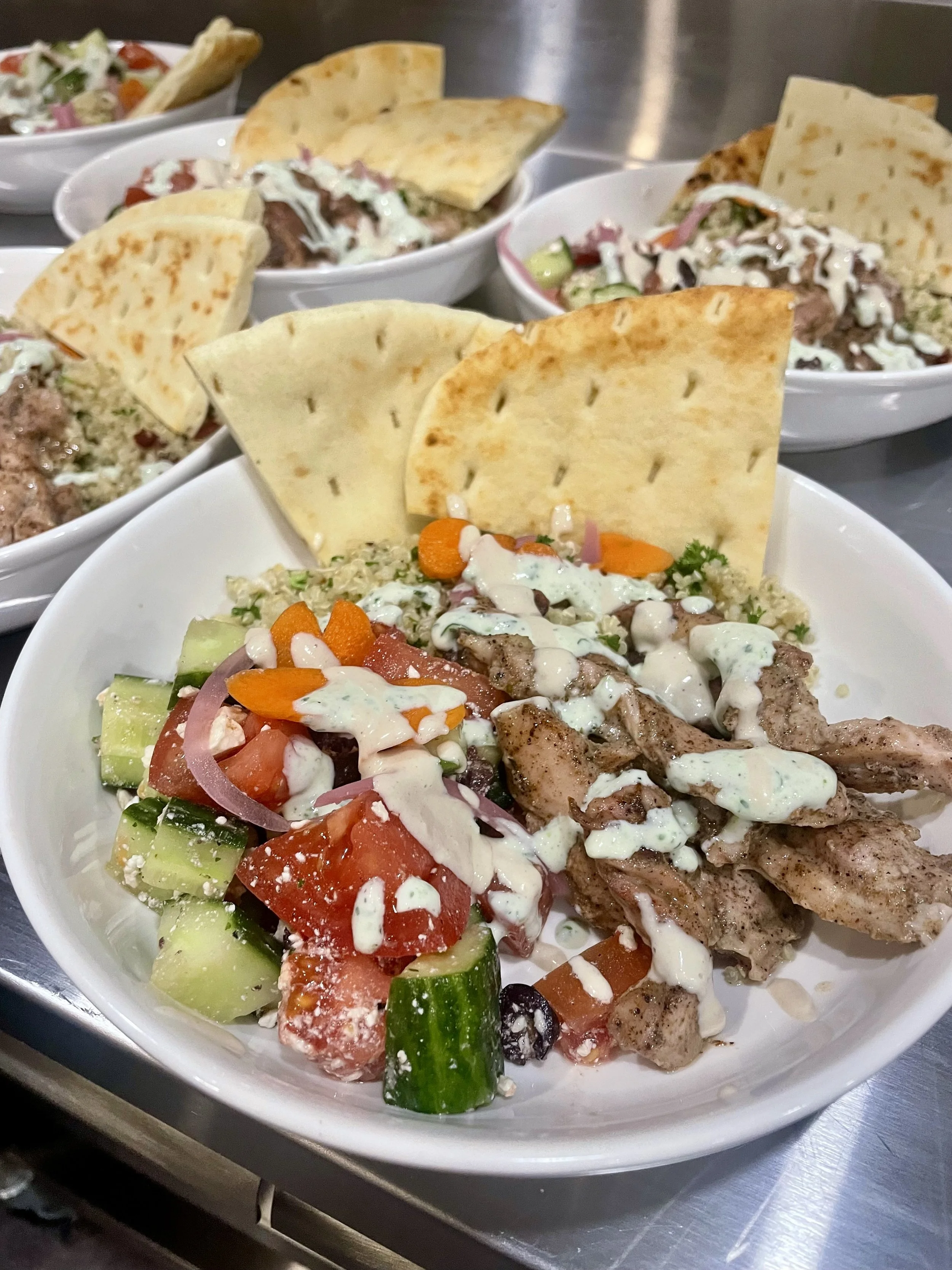 A white bowl filled with salad containing cucumbers, tomatoes, onions, black olives, shredded cheese, chopped herbs, and greens, topped with a creamy dressing, and topped with two pieces of pita prepared by a Revelstoke private chef.
