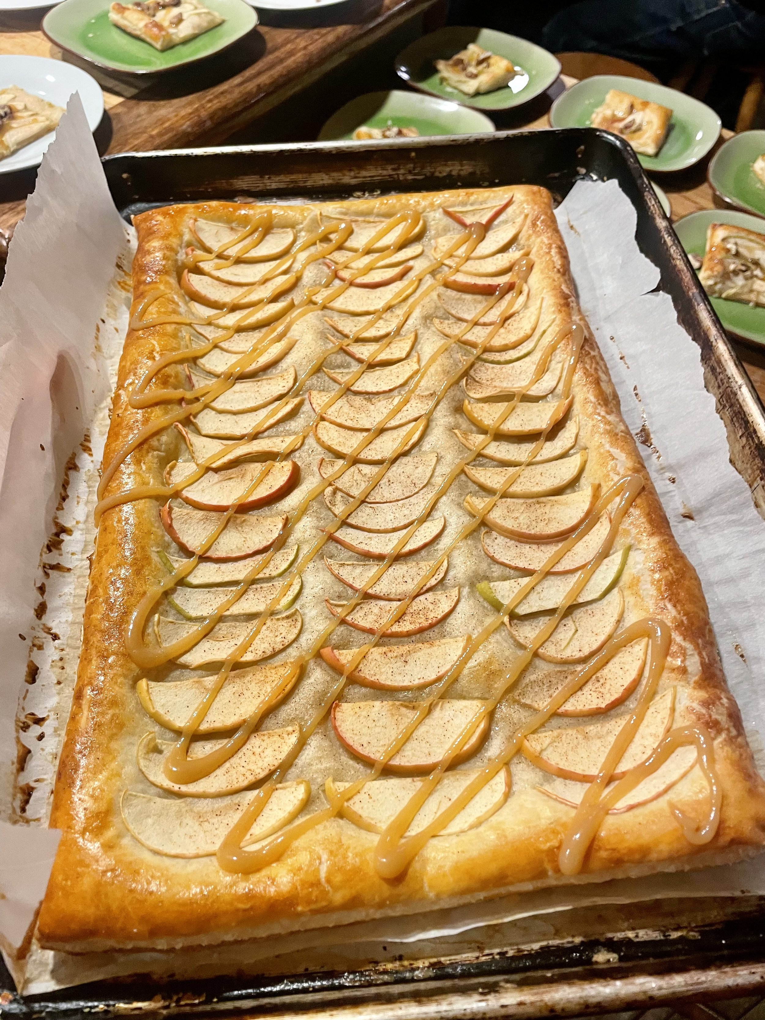 A rectangular fruit tart topped with sliced apples and drizzled caramel sauce, served on a baking tray with parchment paper, surrounded by small green plates with additional slices prepared by a Revelstoke private chef.