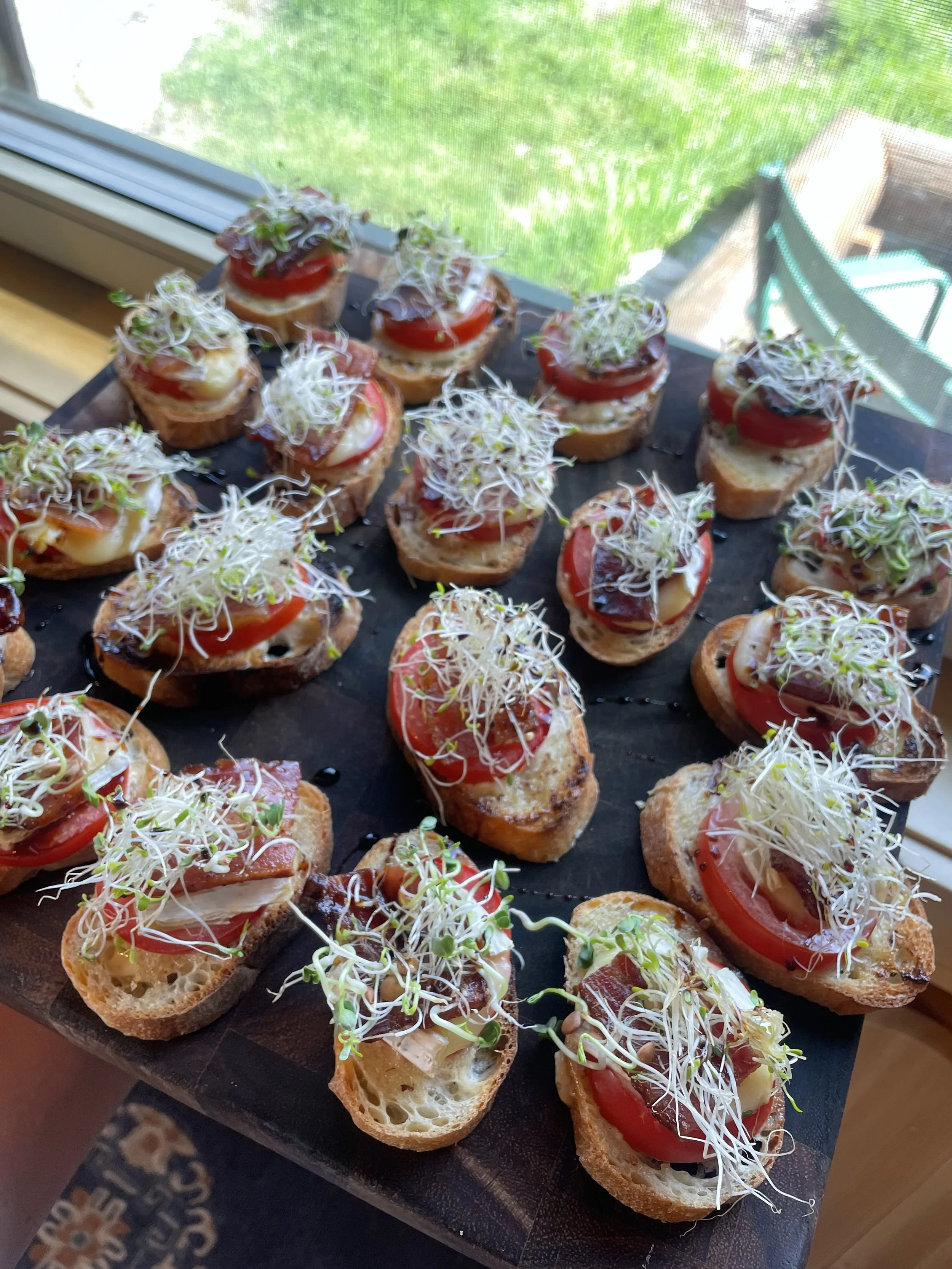 Close-up of sliced bread topped with tomatoes, cheese, cured meat, and microgreens on a black serving board near a window prepared by a Revelstoke private chef.